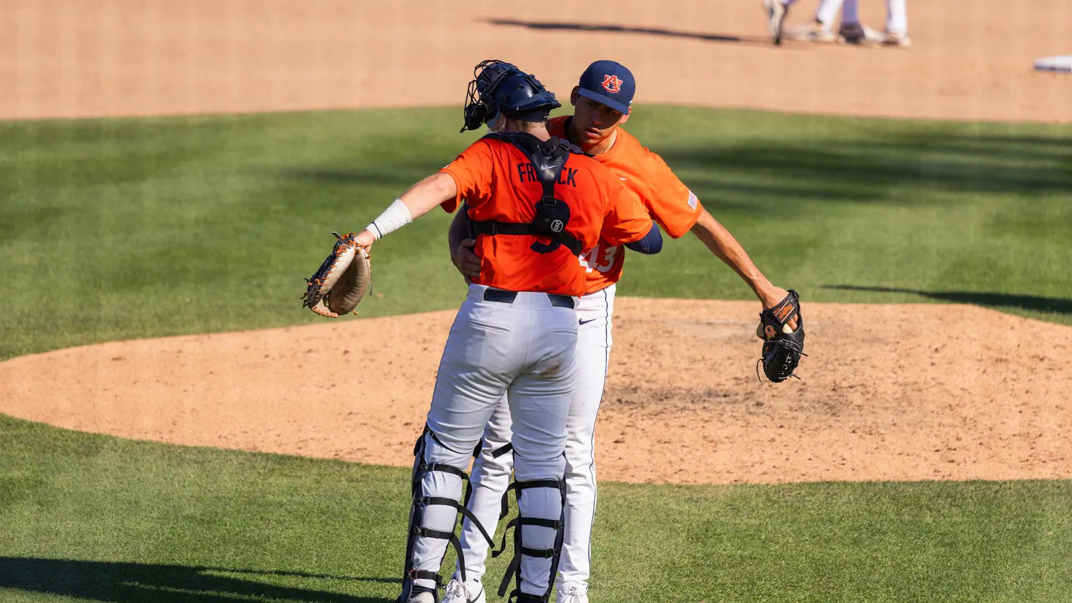 Two baseball players in orange jerseys embrace on the field, with a pitcher in white pants and a catcher wearing gear.