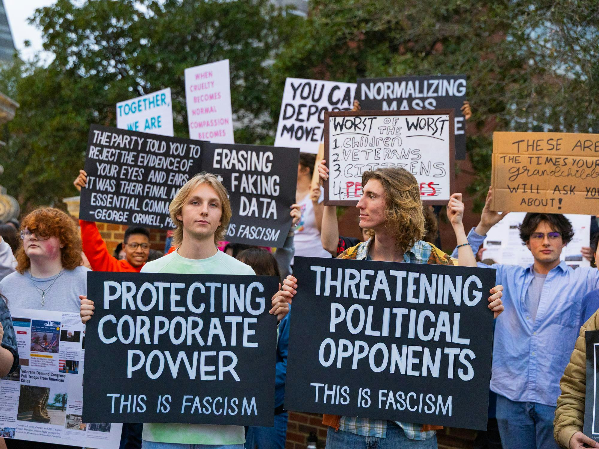 A diverse group of protesters hold signs displaying political messages about corporate power and threats to democracy, amidst a public gathering.