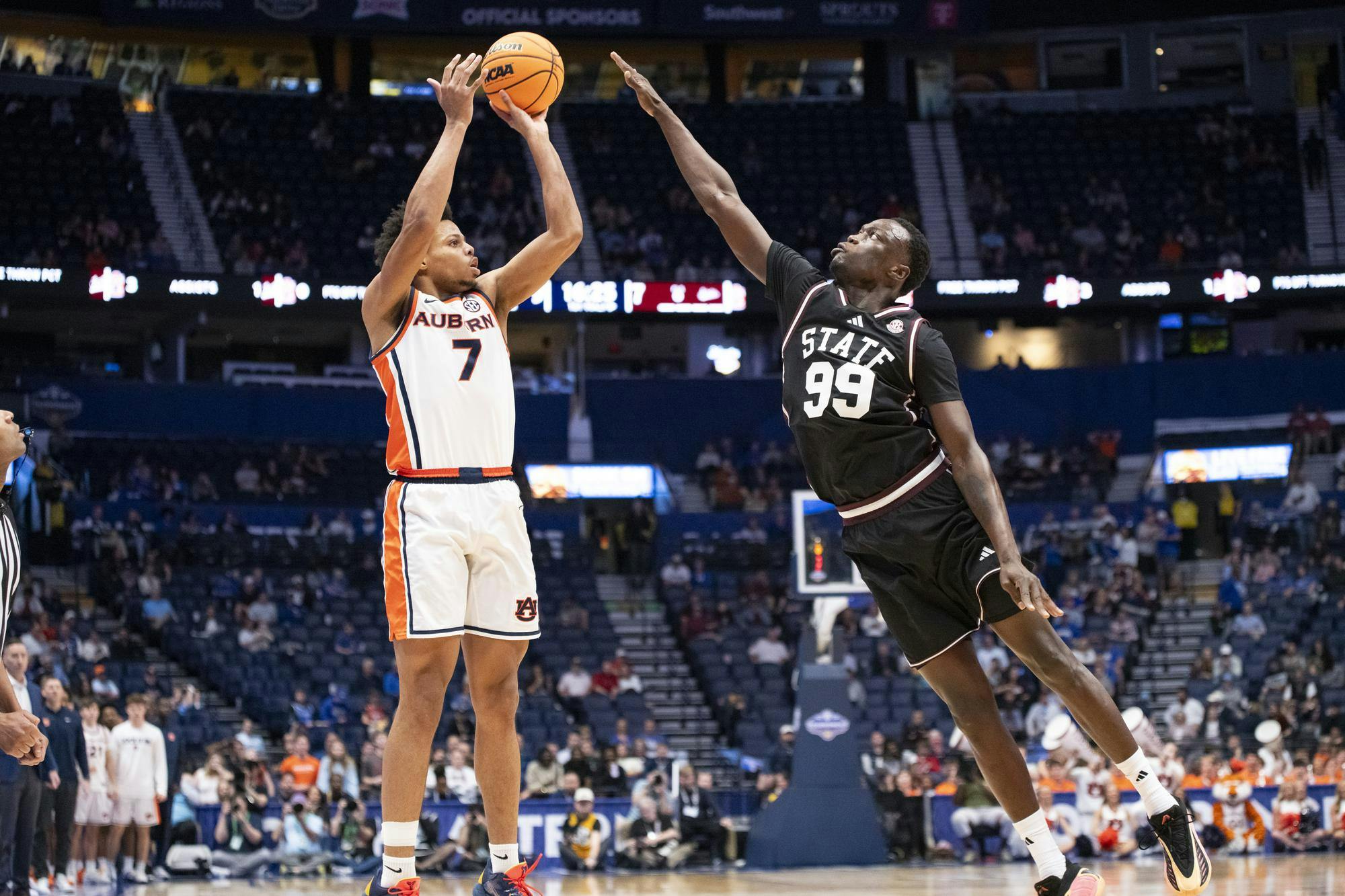 A player in an orange and white uniform shoots a basketball while another player in black jumps to block the shot.