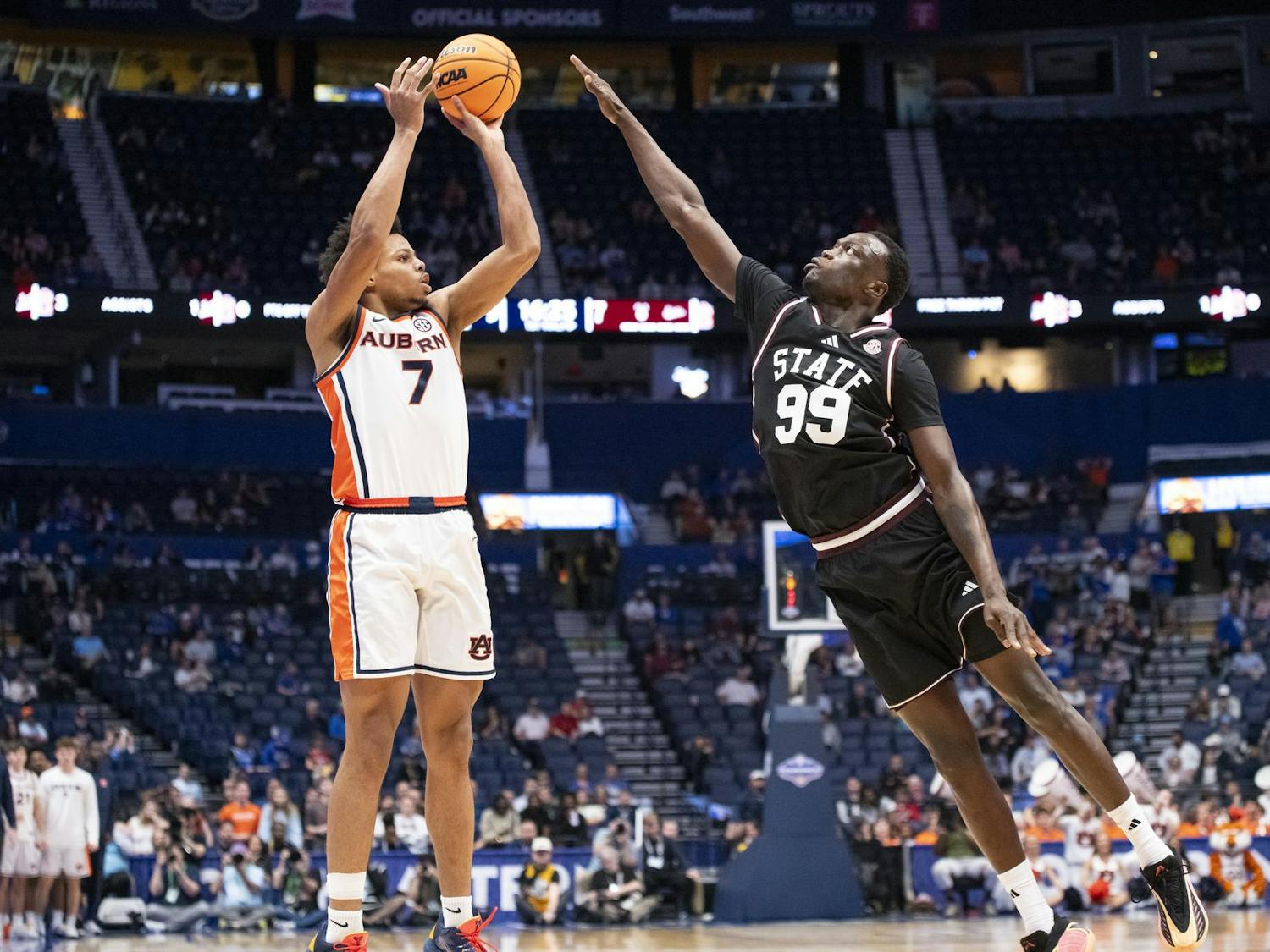 A player in an orange and white uniform shoots a basketball while another player in black jumps to block the shot.