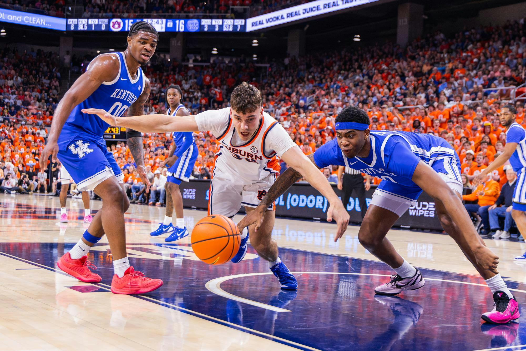 Two players in blue jerseys and one in white are diving for a basketball on a hardwood court, surrounded by an orange-clad crowd.