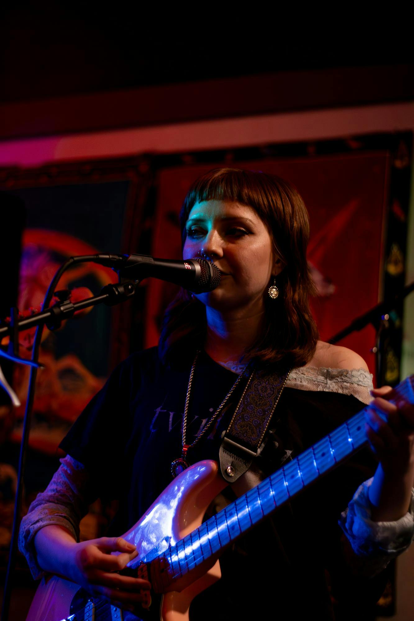A woman with shoulder-length hair sings into a microphone while playing an electric guitar on stage, surrounded by colorful lighting.