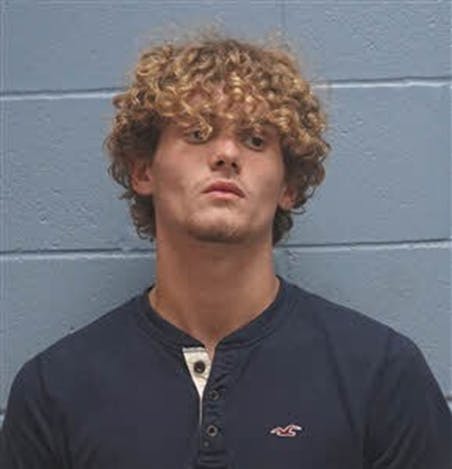 Mugshot of a person with curly hair standing against a light blue wall, wearing a dark shirt with a buttoned neckline.
