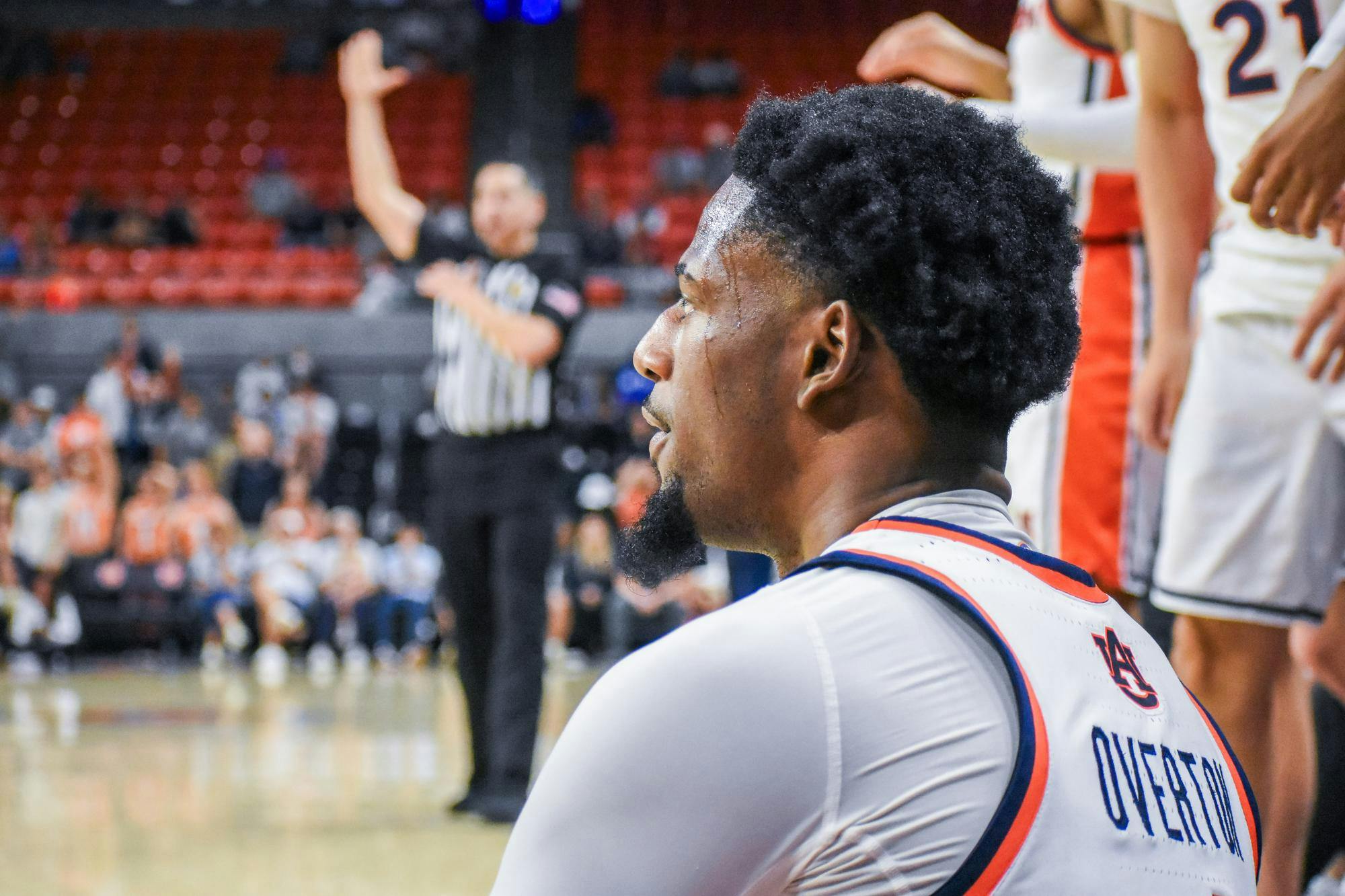 A player with a closely cropped hairstyle sits on the sidelines, while a referee signals with one hand raised in the background.