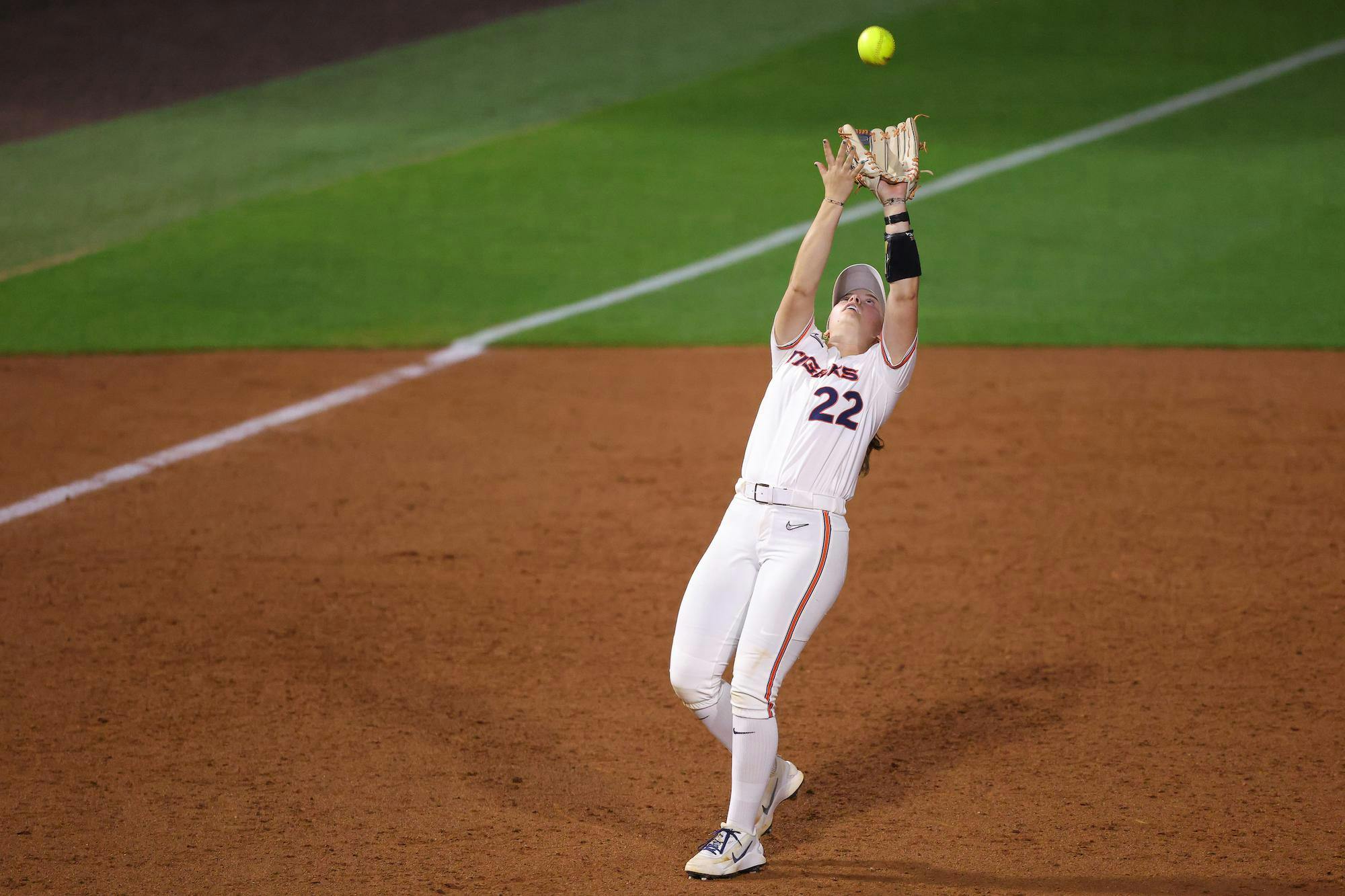 A player in white uniform reaches high to catch a flying yellow softball during a game on a dirt field.