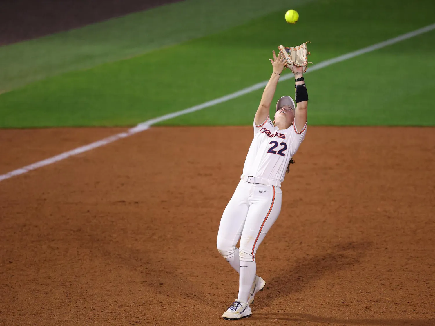 A player in white uniform reaches high to catch a flying yellow softball during a game on a dirt field.