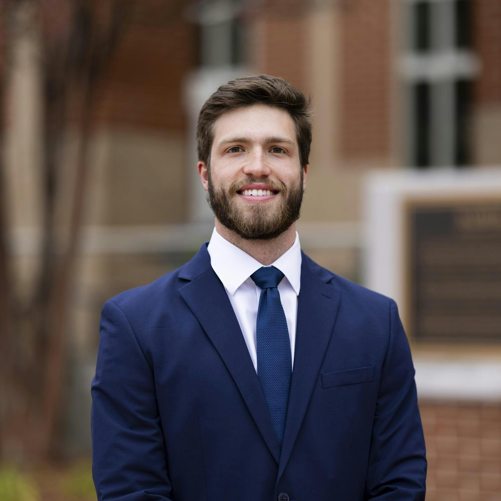 A man with a beard smiles while wearing a dark suit, white shirt, and tie, standing outdoors with a blurred background.