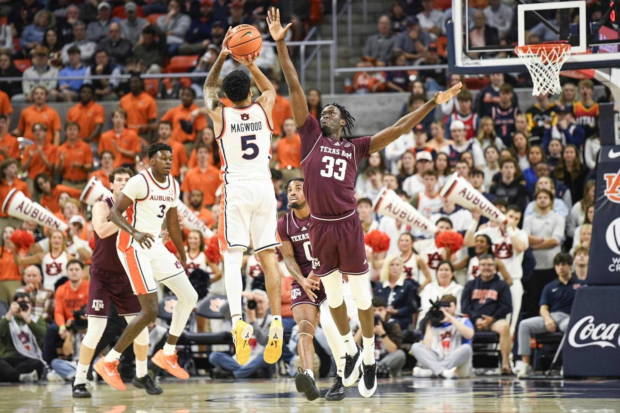 A basketball player in a white jersey jumps to shoot, while opponents in maroon try to block, amidst an energetic crowd.