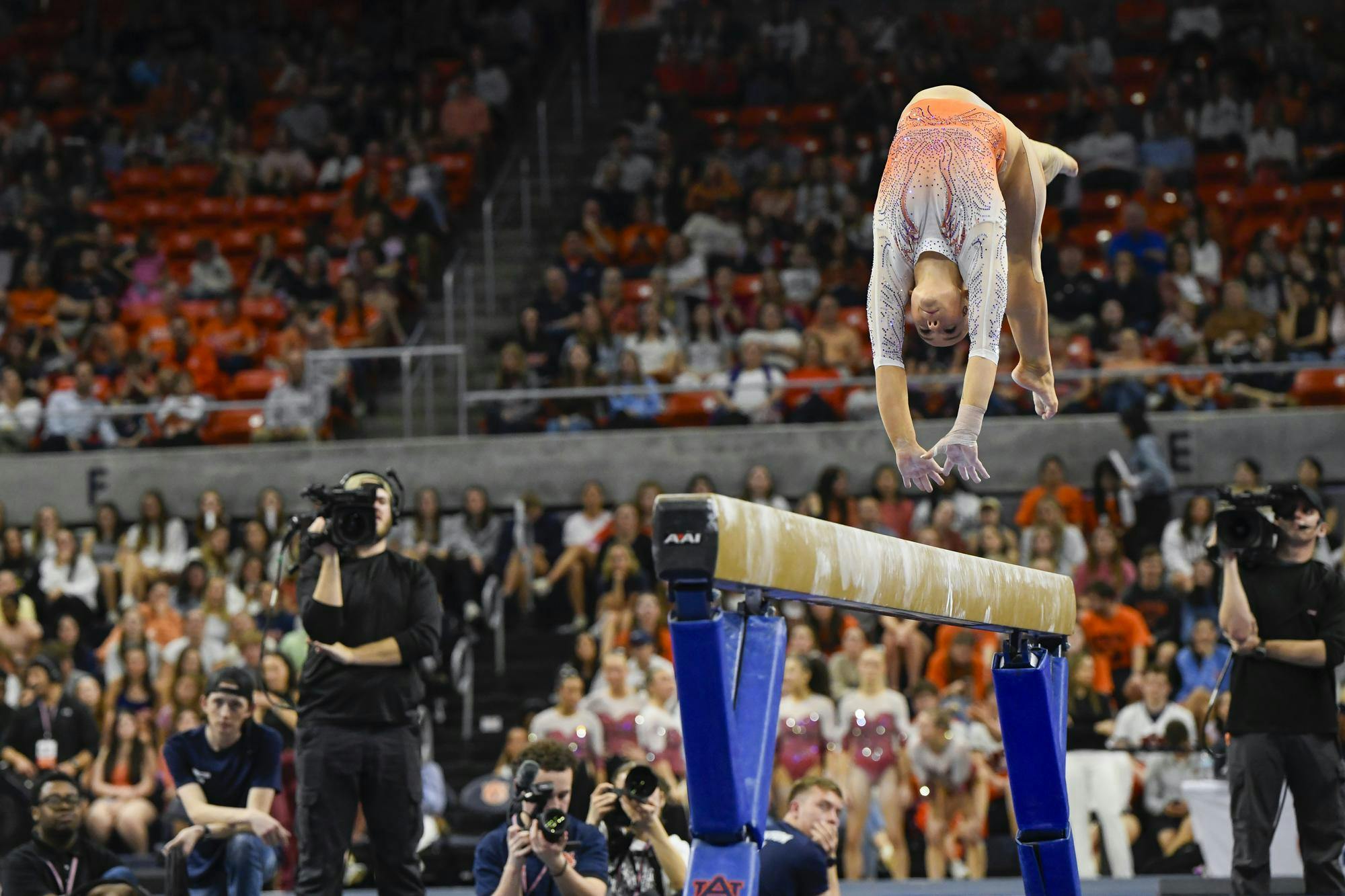 A gymnast is performing an aerial maneuver above a balance beam while photographers capture the action in a crowded arena.