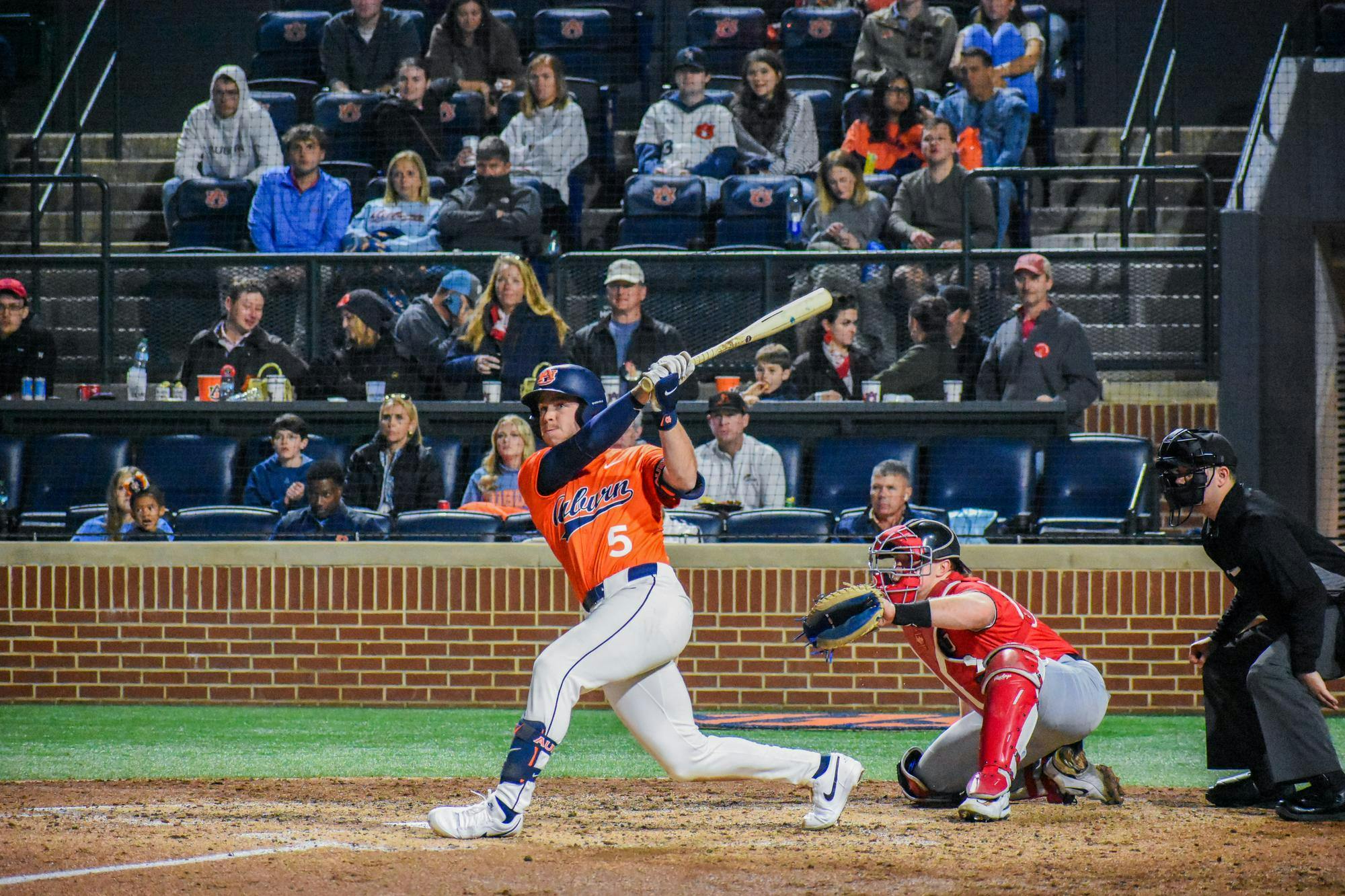 A baseball player swings a bat as a catcher crouches behind him, surrounded by a crowd in the stands.