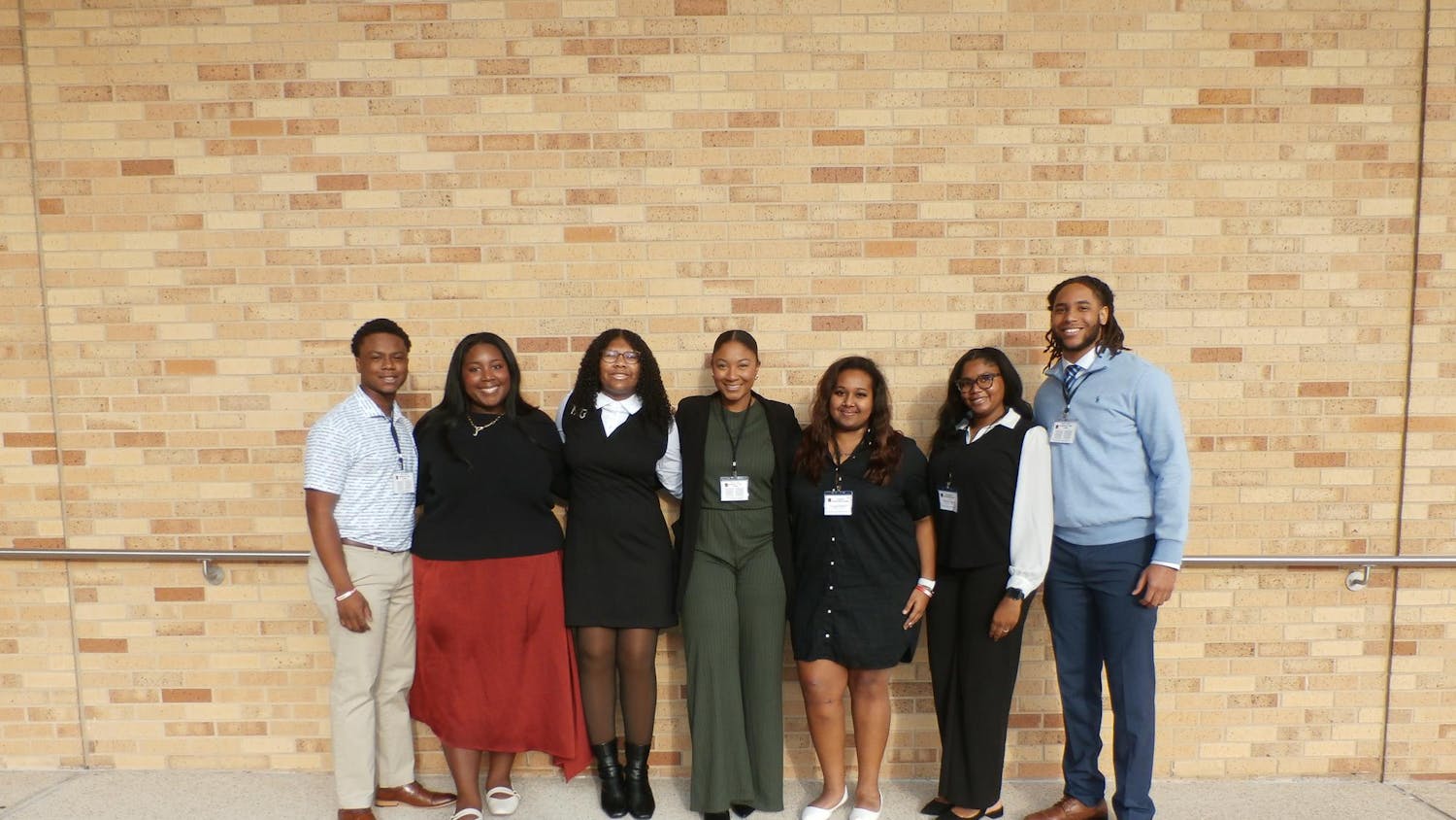 Seven individuals stand in a row against a brick wall, wearing formal attire and name tags.