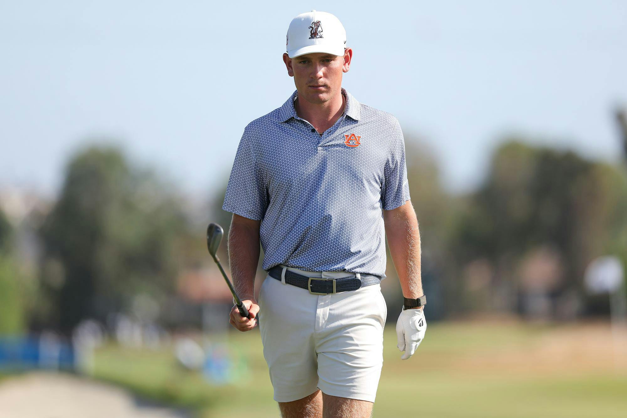 A young golfer walks on a golf course, holding a club, wearing a shirt with a logo and white shorts.