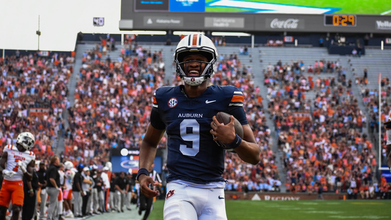 A football player in a navy jersey with an auburn logo runs joyfully on a field, holding a football, while a crowd cheers.
