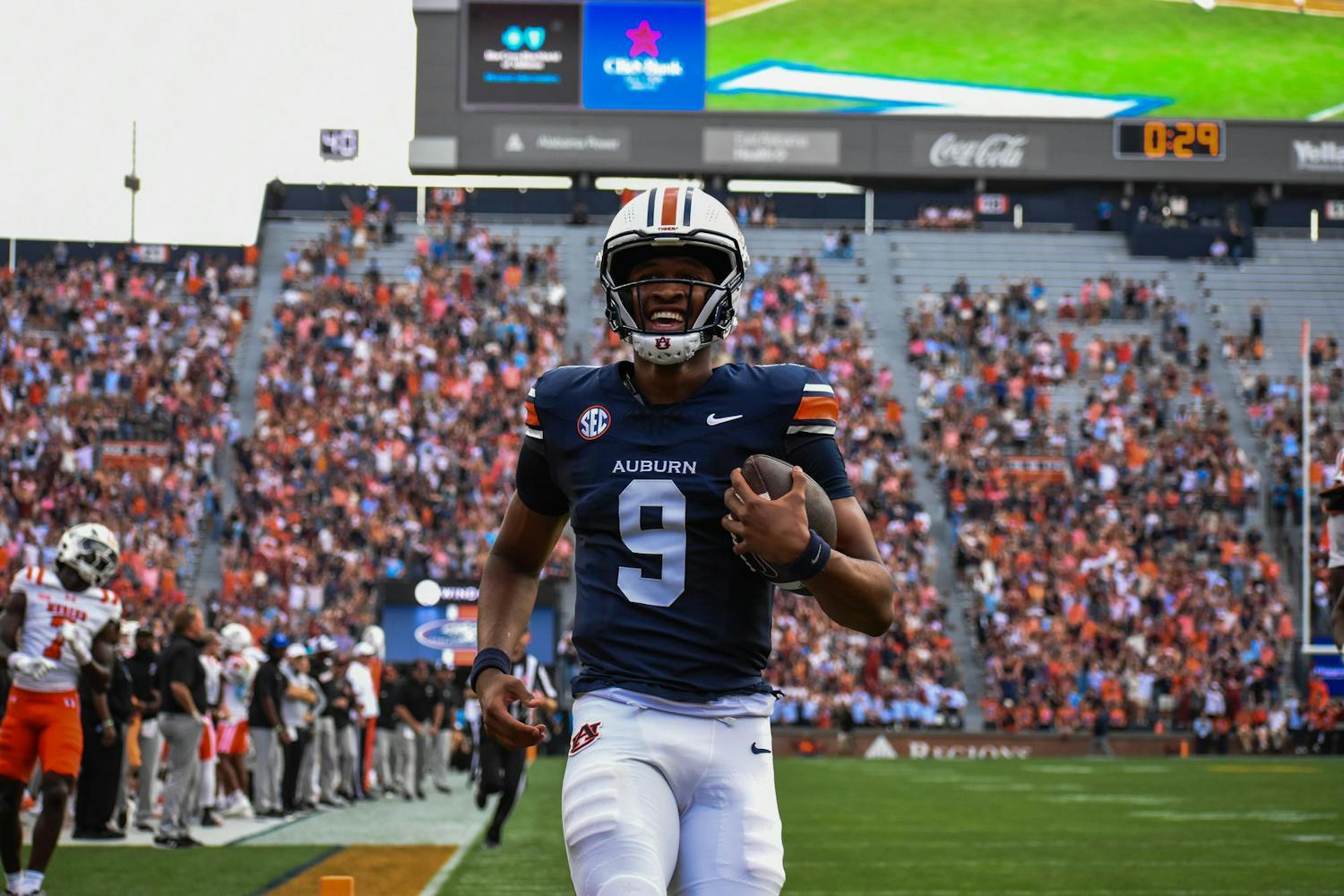 A football player in a navy jersey with an auburn logo runs joyfully on a field, holding a football, while a crowd cheers.
