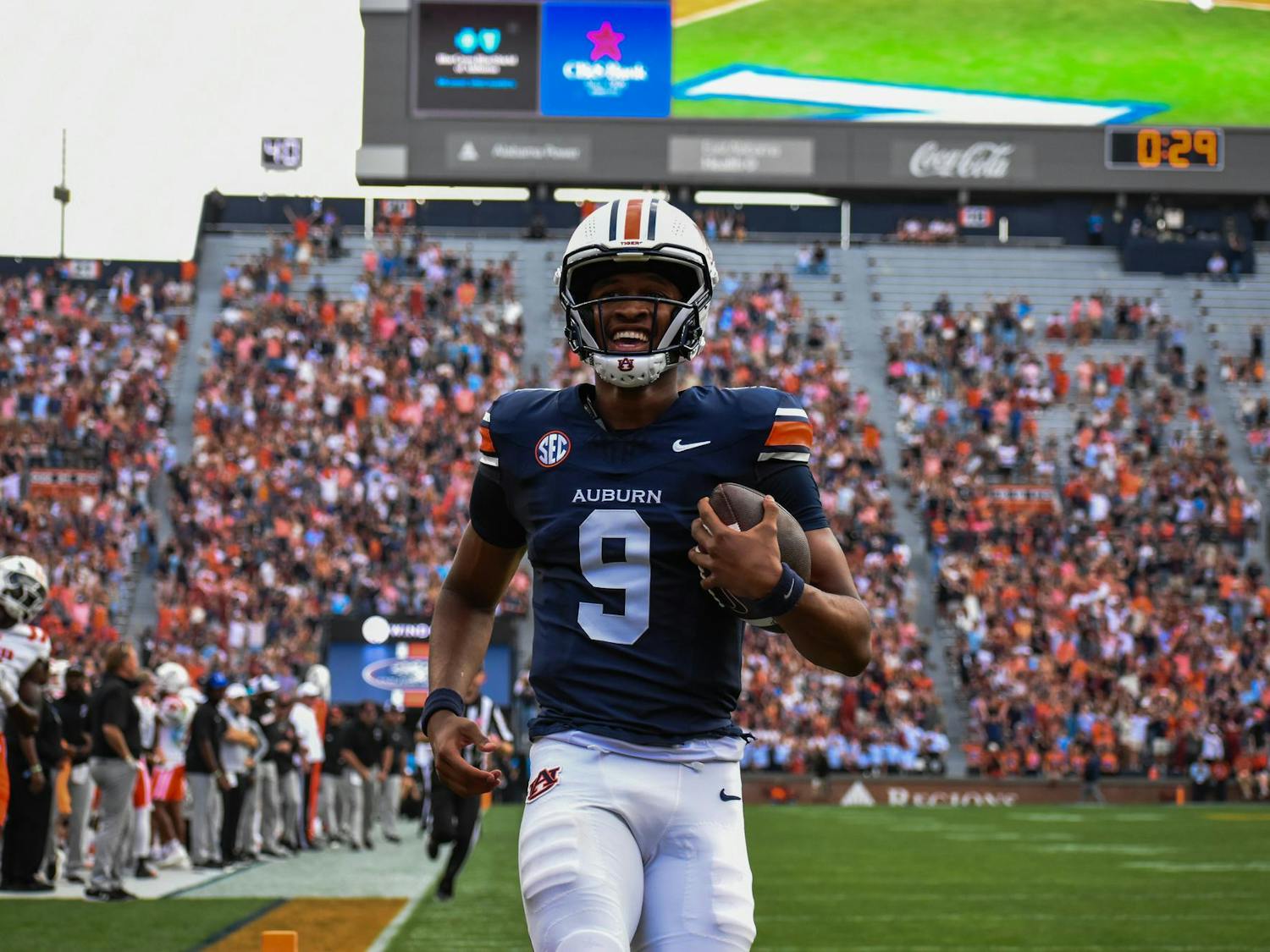 A football player in a navy jersey with an auburn logo runs joyfully on a field, holding a football, while a crowd cheers.
