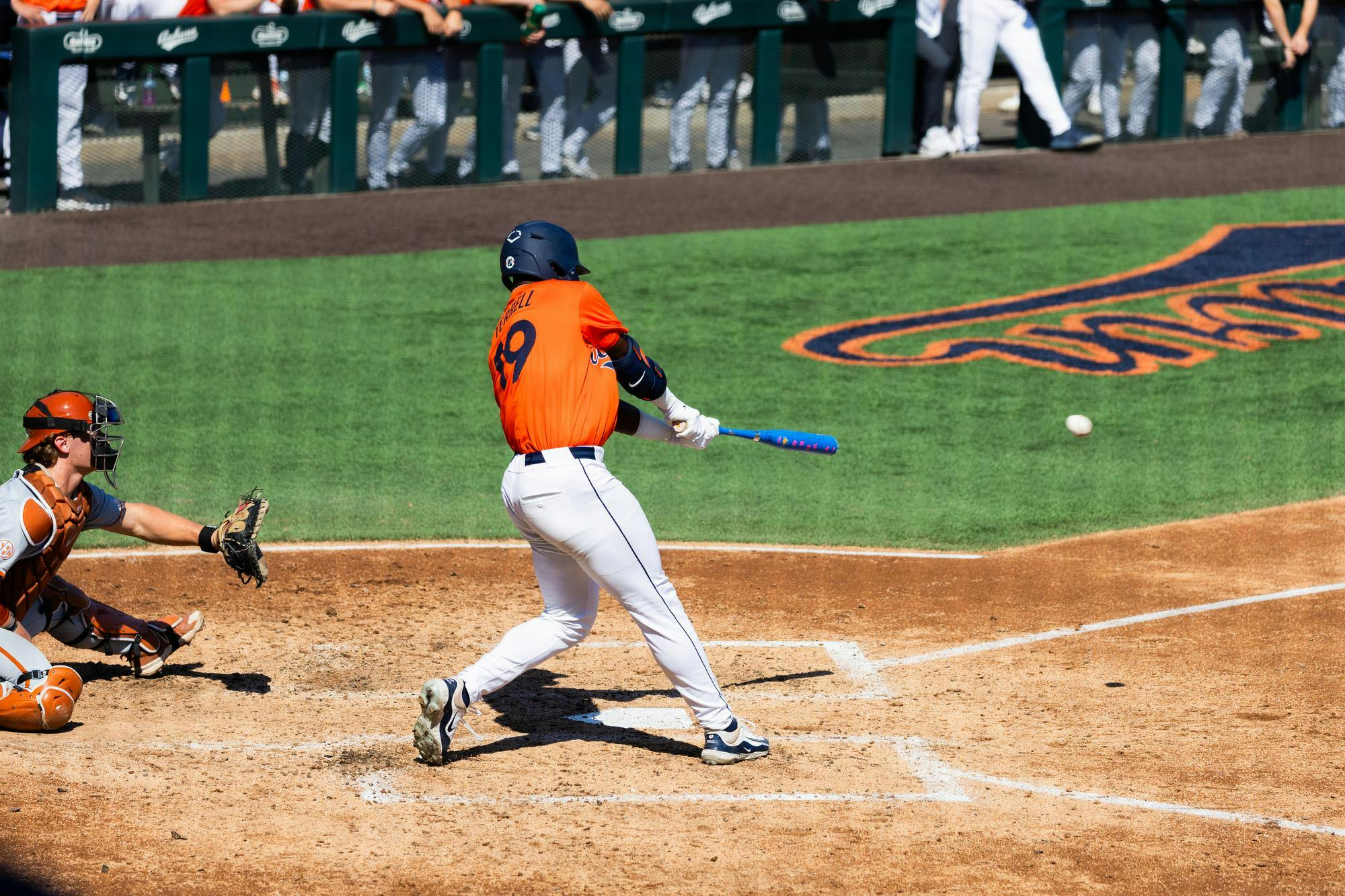 A batter in an orange jersey swings a blue bat at a baseball, while a catcher in gray prepares to receive it.
