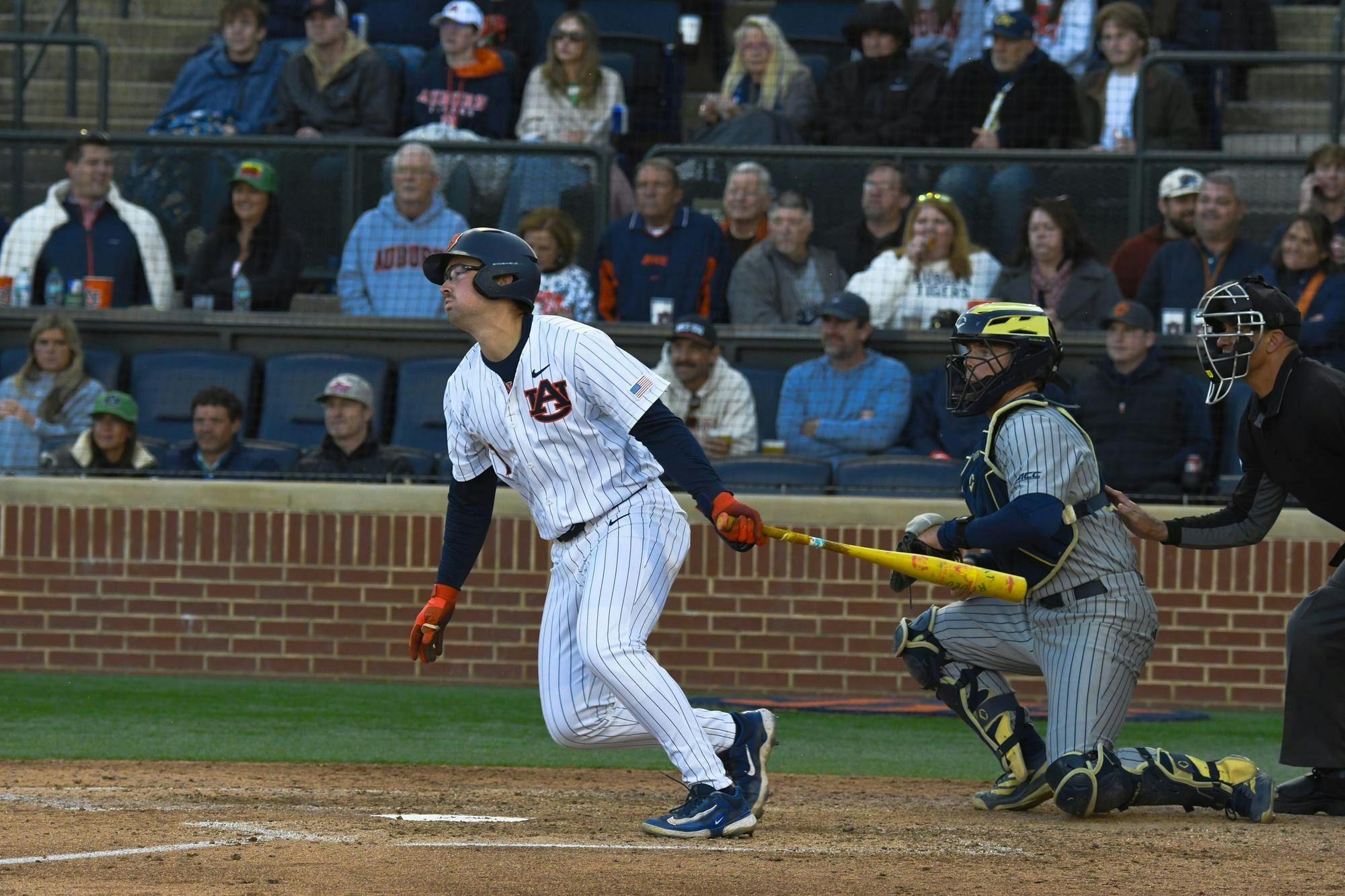 A baseball player in a white striped uniform swings a bat while a catcher crouches behind him, with an audience in the background.