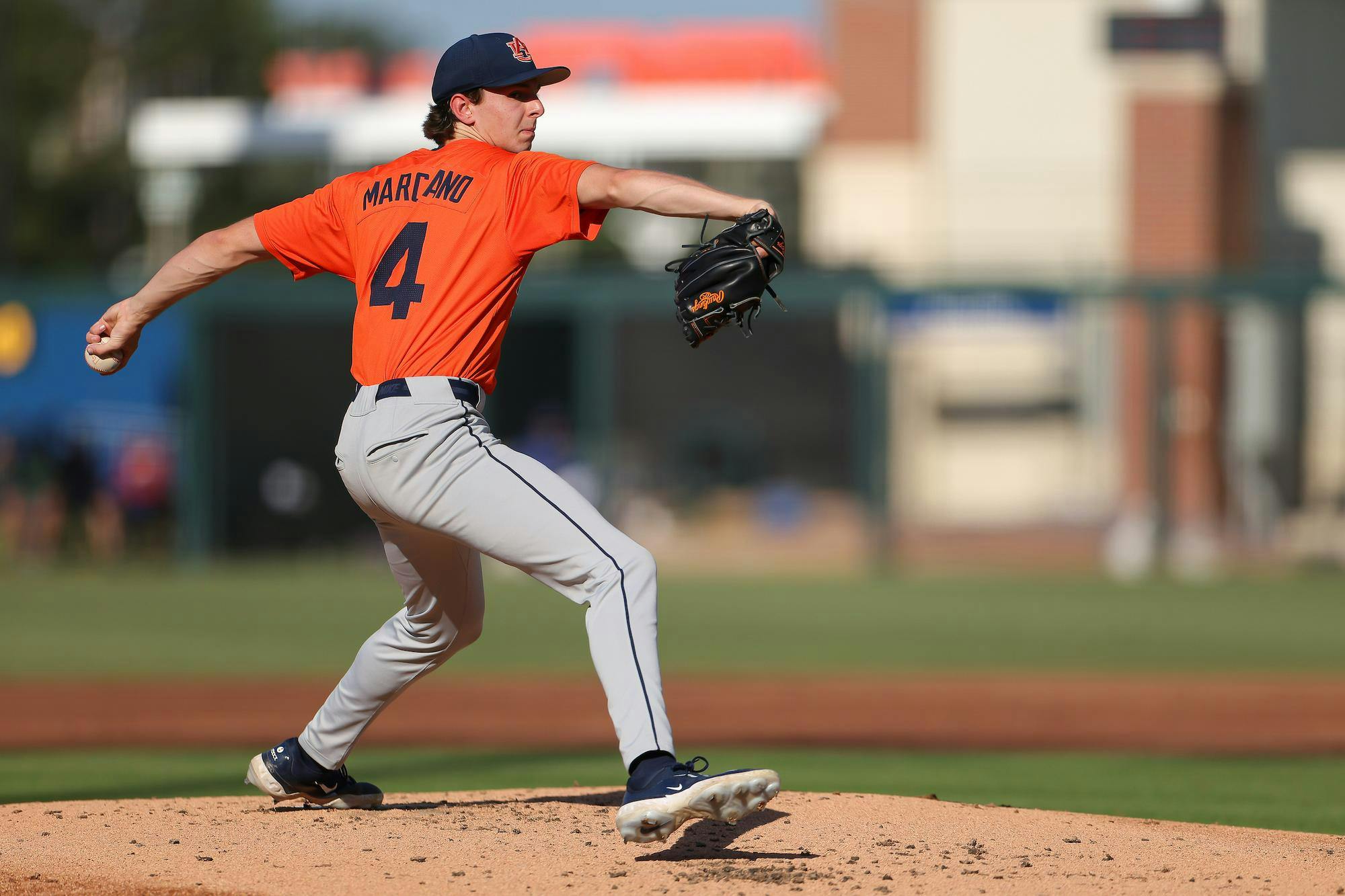 A baseball pitcher in an orange jersey and gray pants delivers a pitch from the mound, focused and poised.