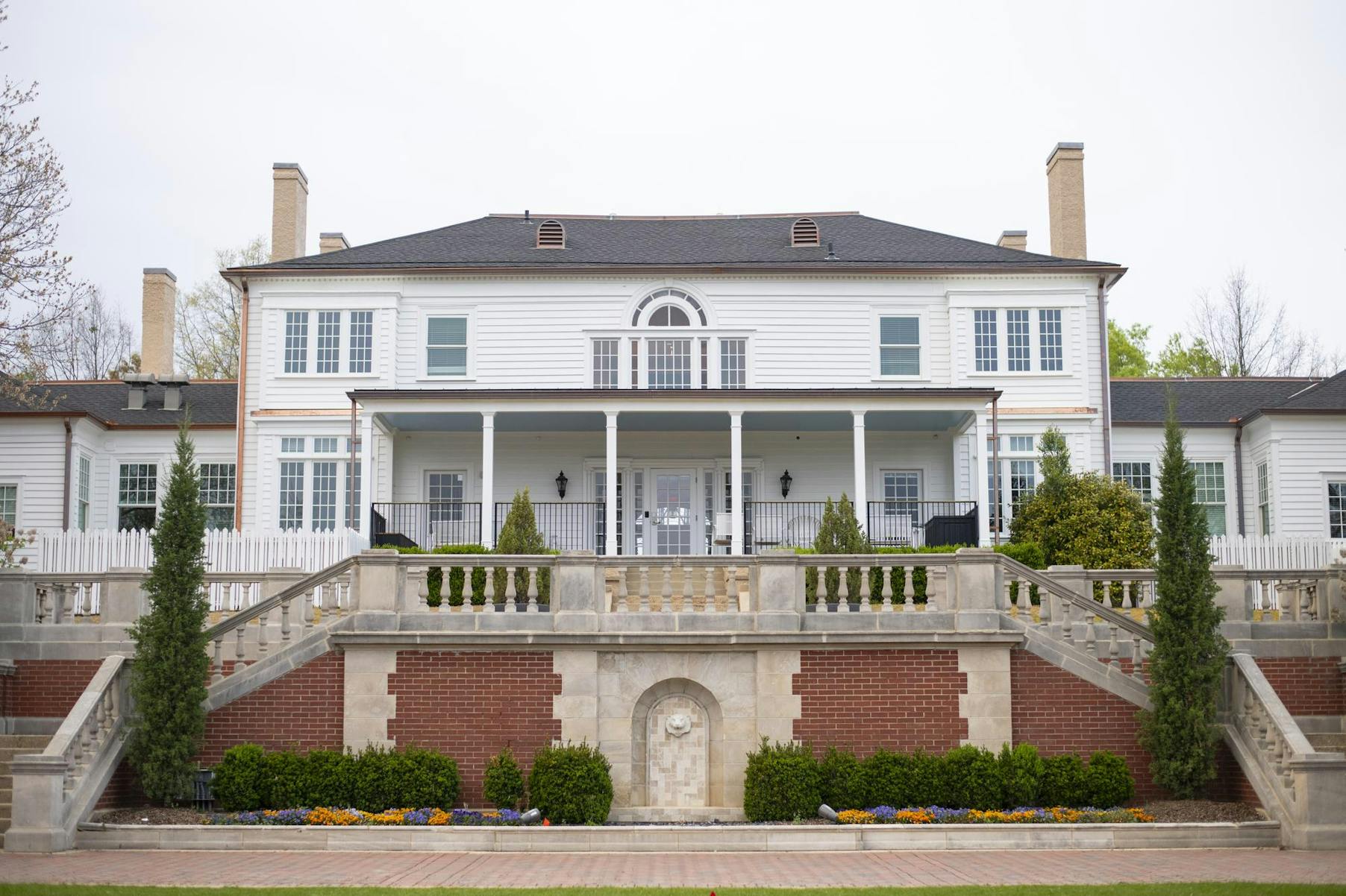 A large white mansion with multiple windows features a wide staircase leading to a porch and landscaped gardens below.