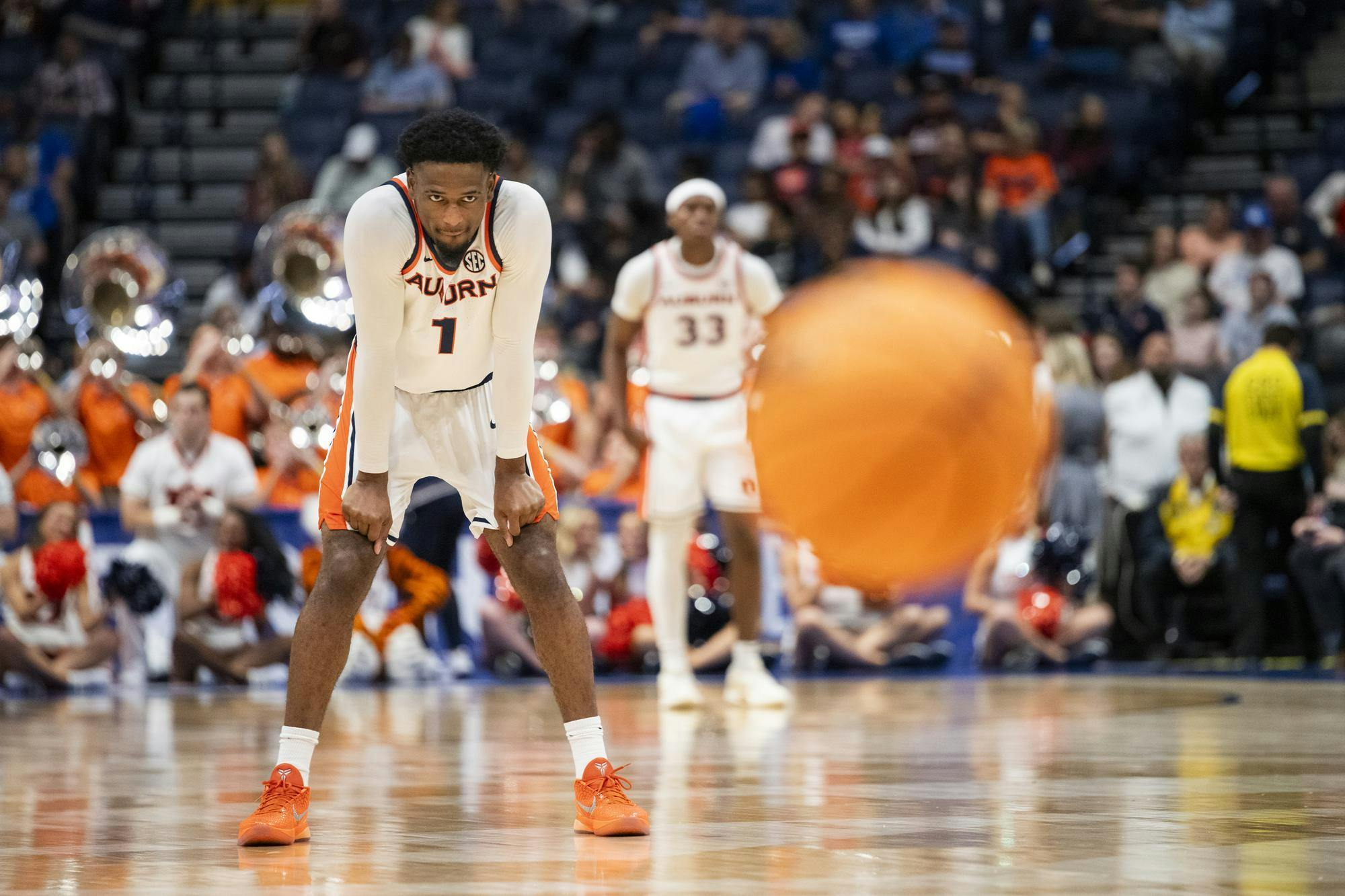 A basketball player in an orange and white uniform stands focused with a large basketball blurred in motion toward him, surrounded by cheering fans and teammates.