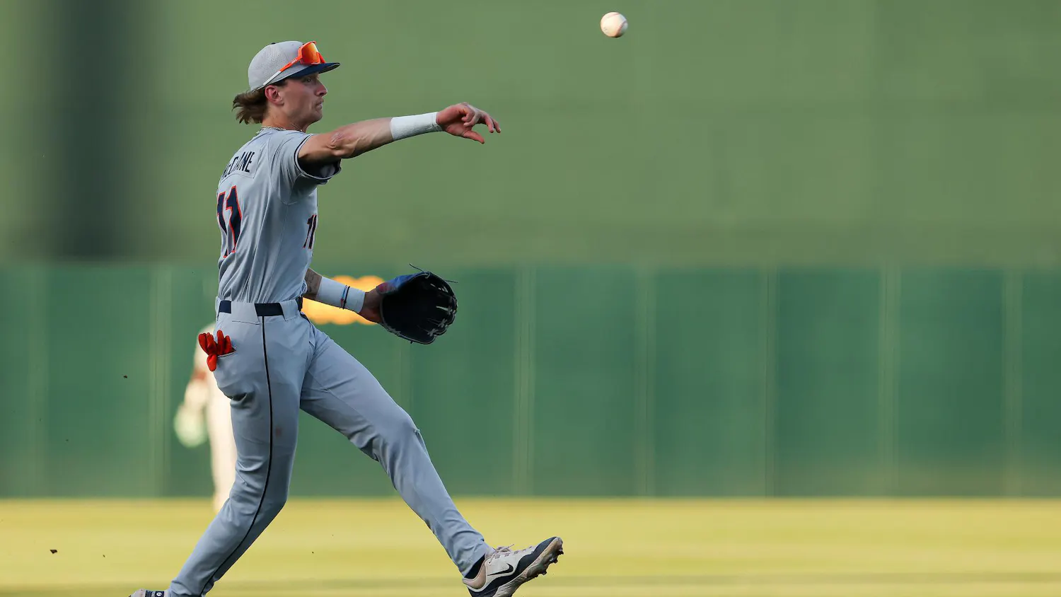 A baseball player in a gray uniform leaps to throw a ball while standing on a dirt infield.