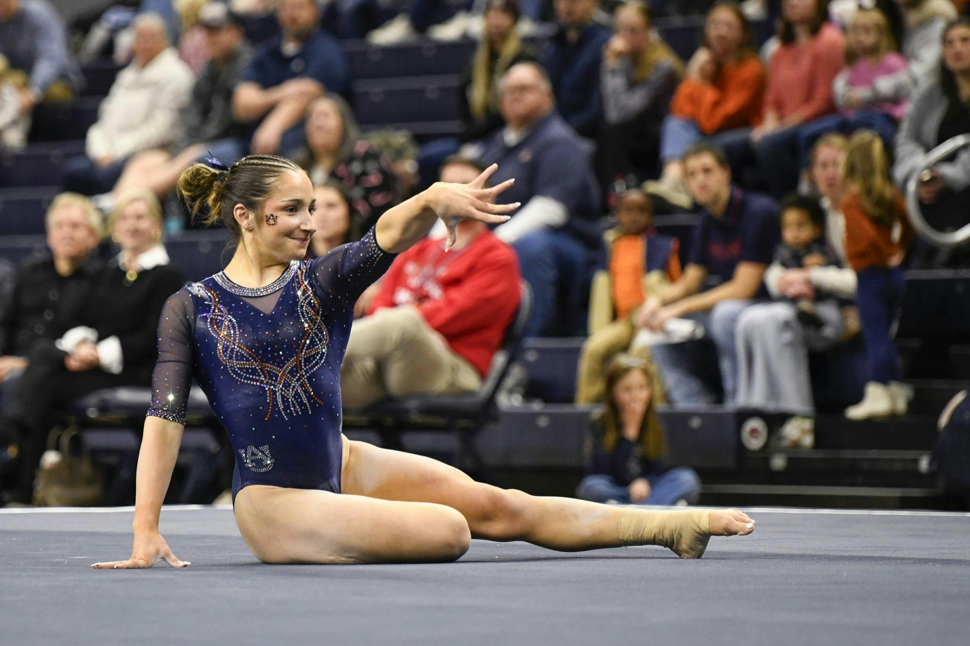 A gymnast in a decorated blue leotard performs on a mat, while spectators watch in the background.