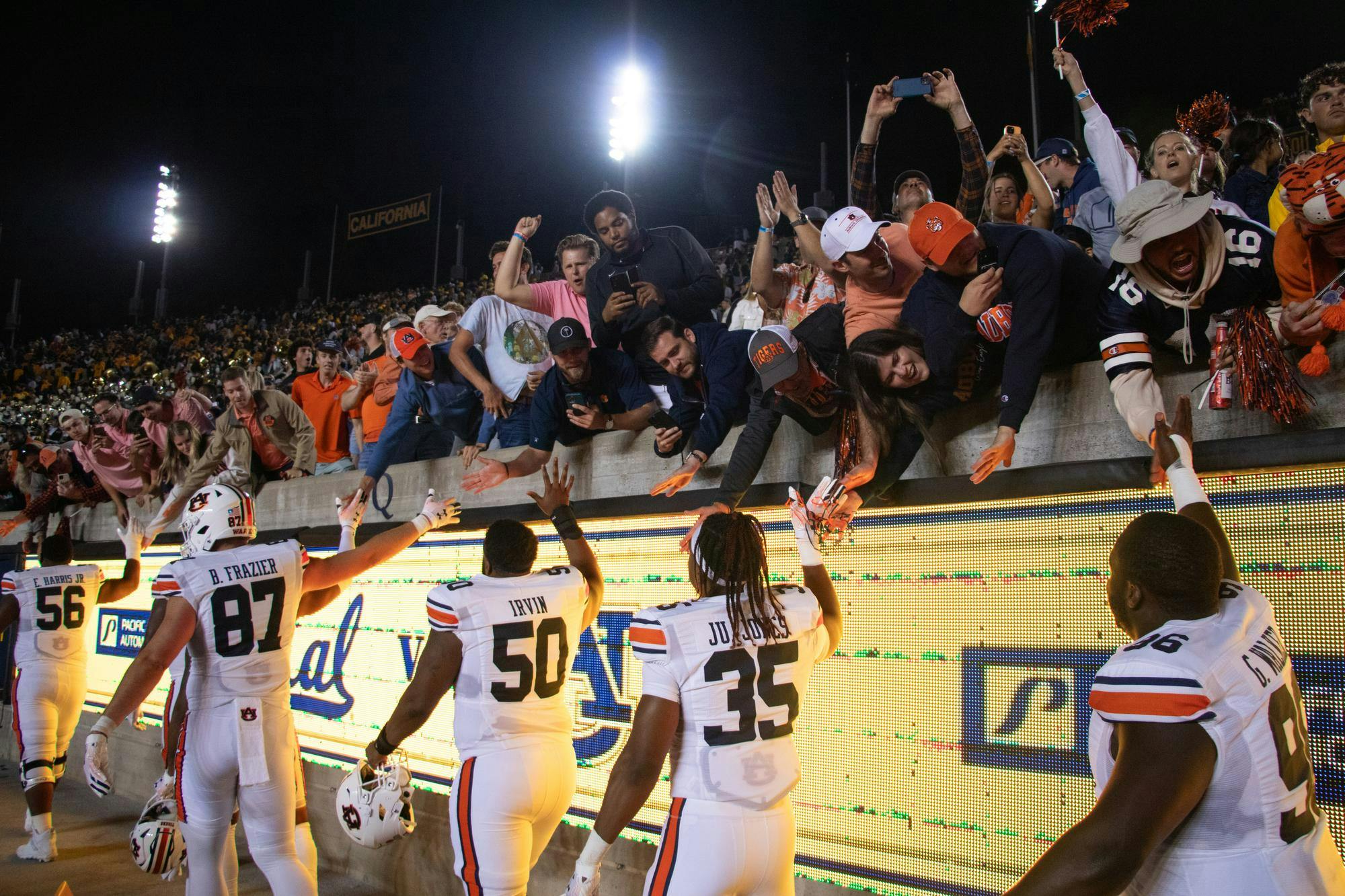 Football players in white jerseys interact with enthusiastic fans reaching over a barrier, celebrating a victorious moment in a stadium at night.