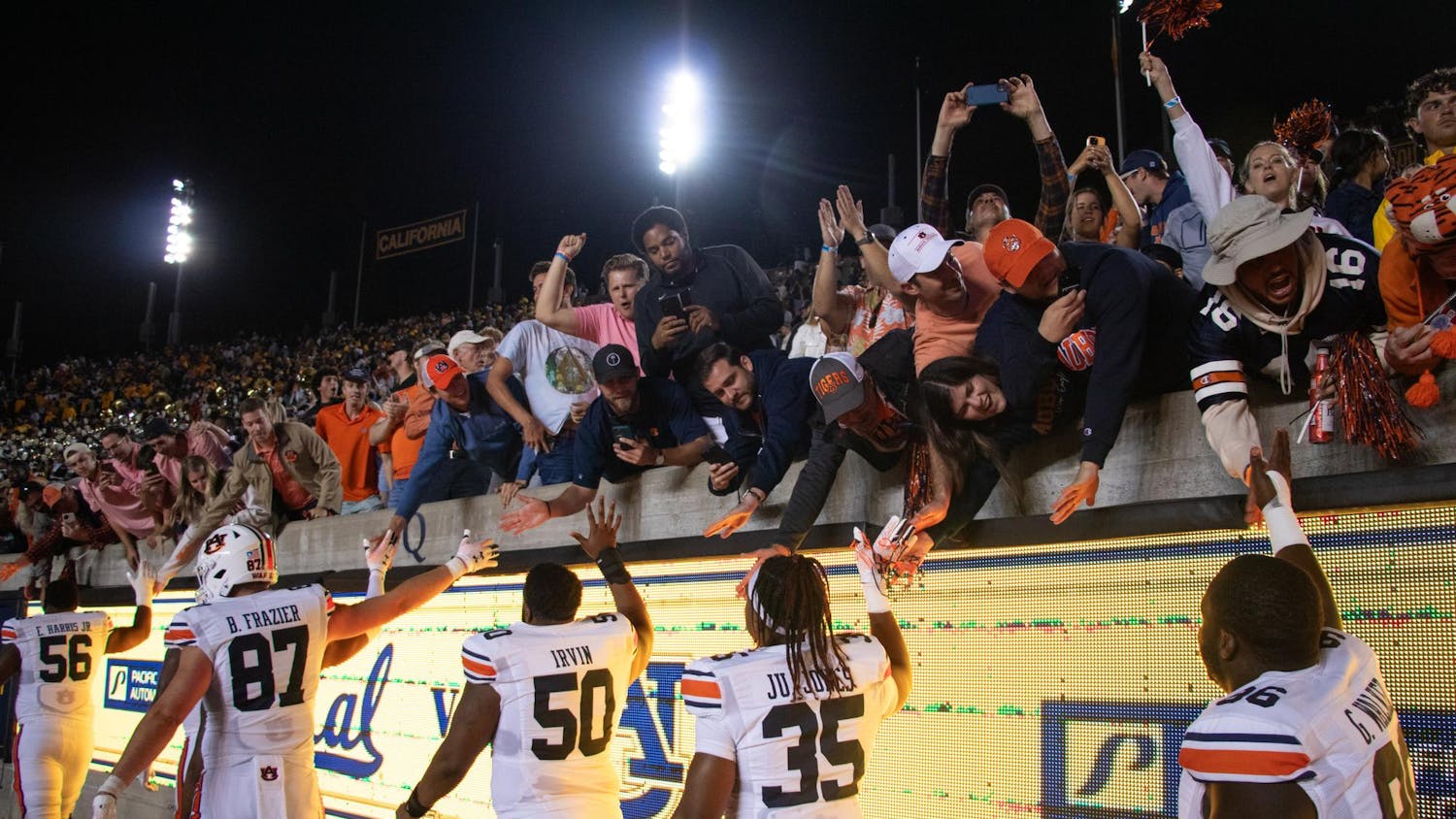 Football players in white jerseys interact with enthusiastic fans reaching over a barrier, celebrating a victorious moment in a stadium at night.
