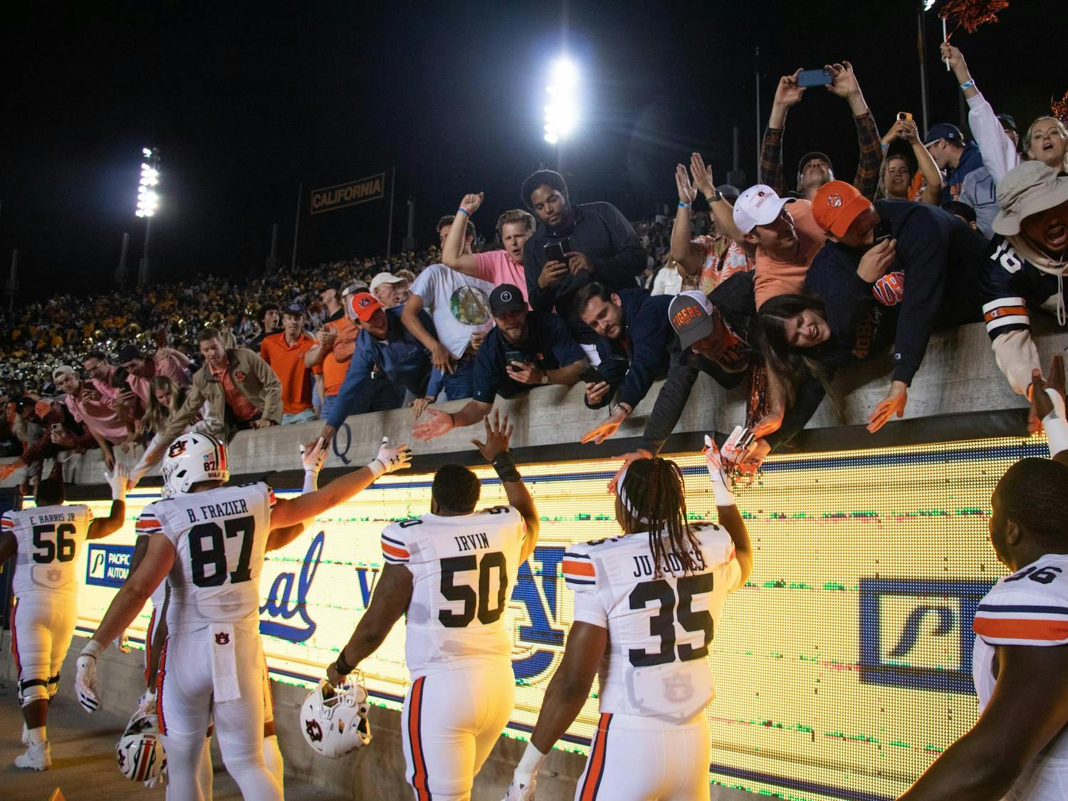 Football players in white jerseys interact with enthusiastic fans reaching over a barrier, celebrating a victorious moment in a stadium at night.