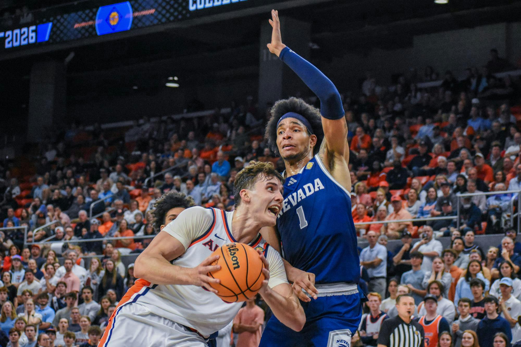 A player in a white jersey fiercely grips a basketball while another player in blue reaches to block him, amidst a cheering crowd.