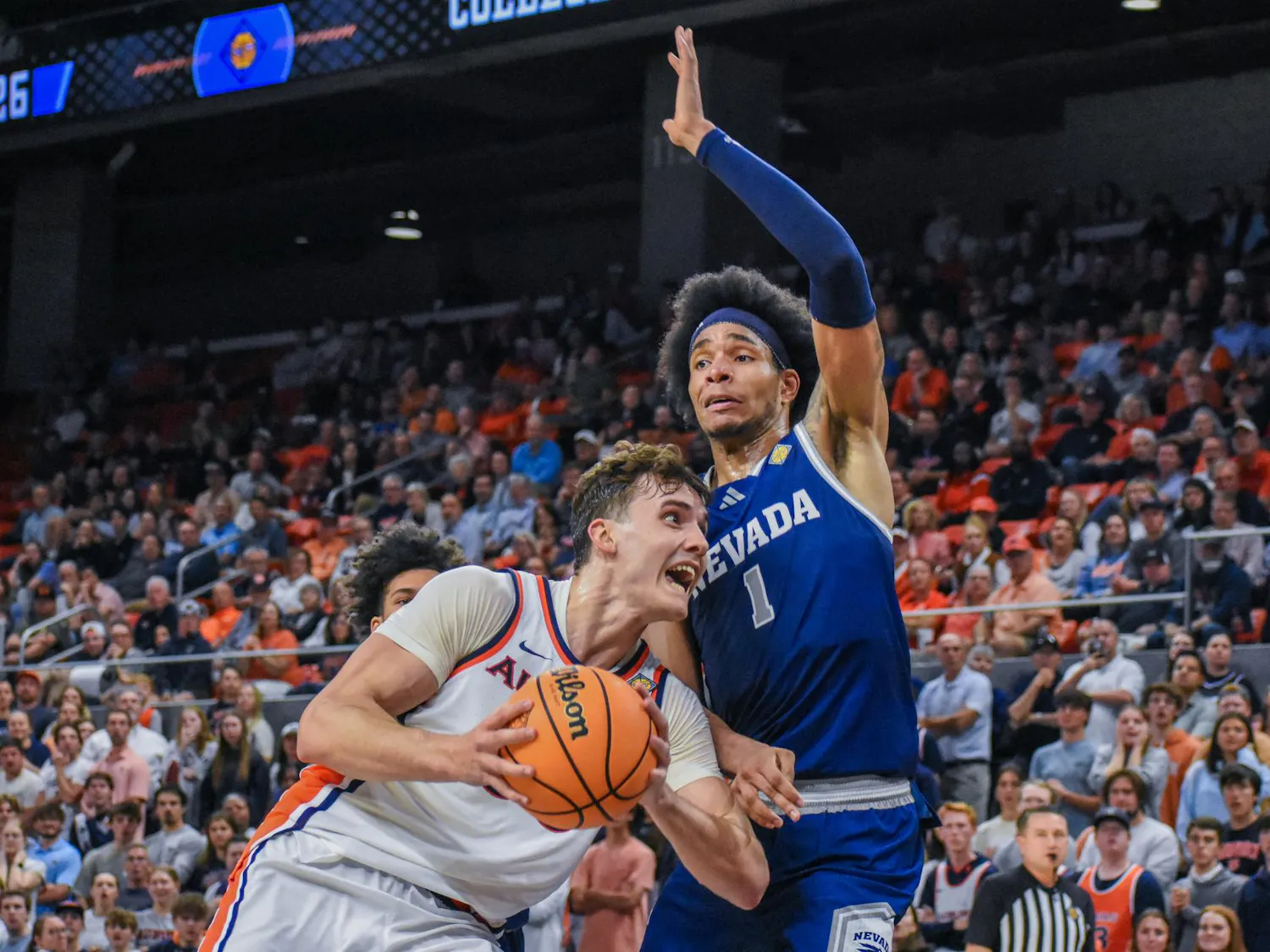A player in a white jersey fiercely grips a basketball while another player in blue reaches to block him, amidst a cheering crowd.