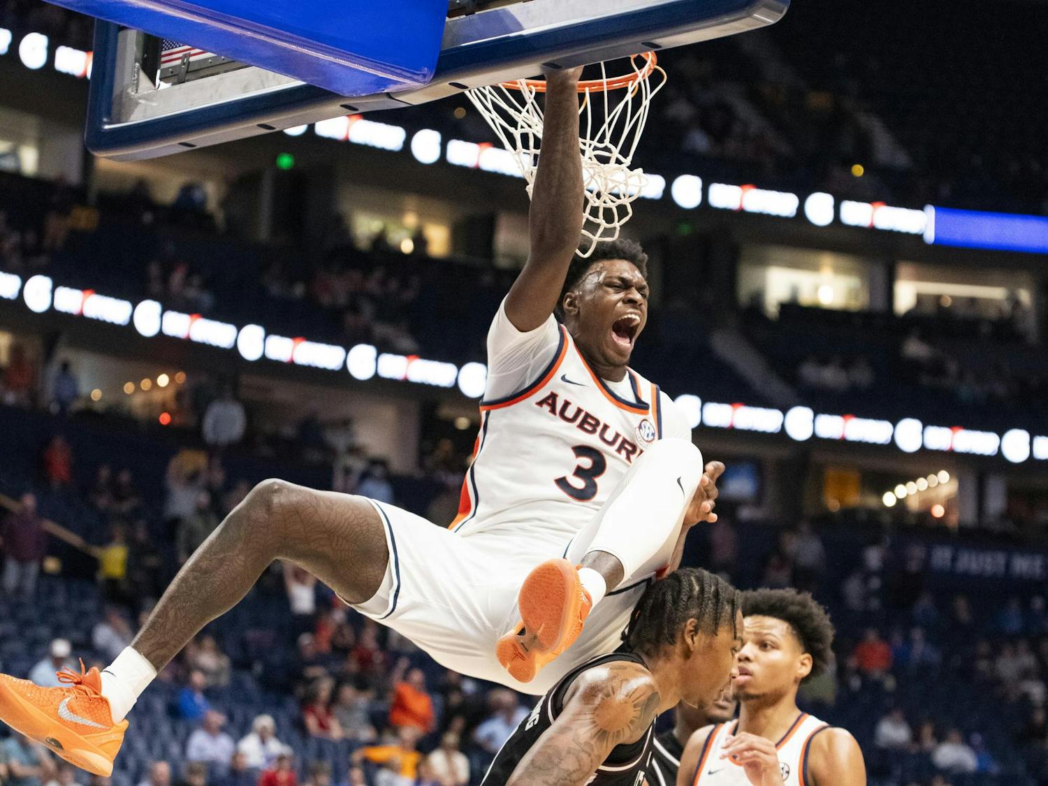 A basketball player in an Auburn jersey hangs from the hoop, celebrating while two opponents look on.