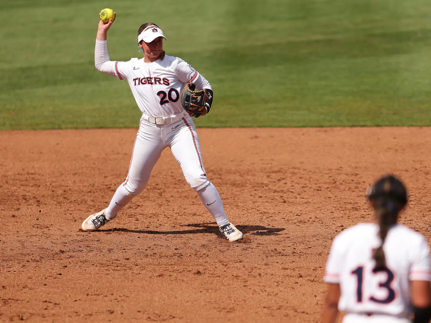 A player in a white uniform throws a yellow softball while another player in a similar uniform stands nearby.