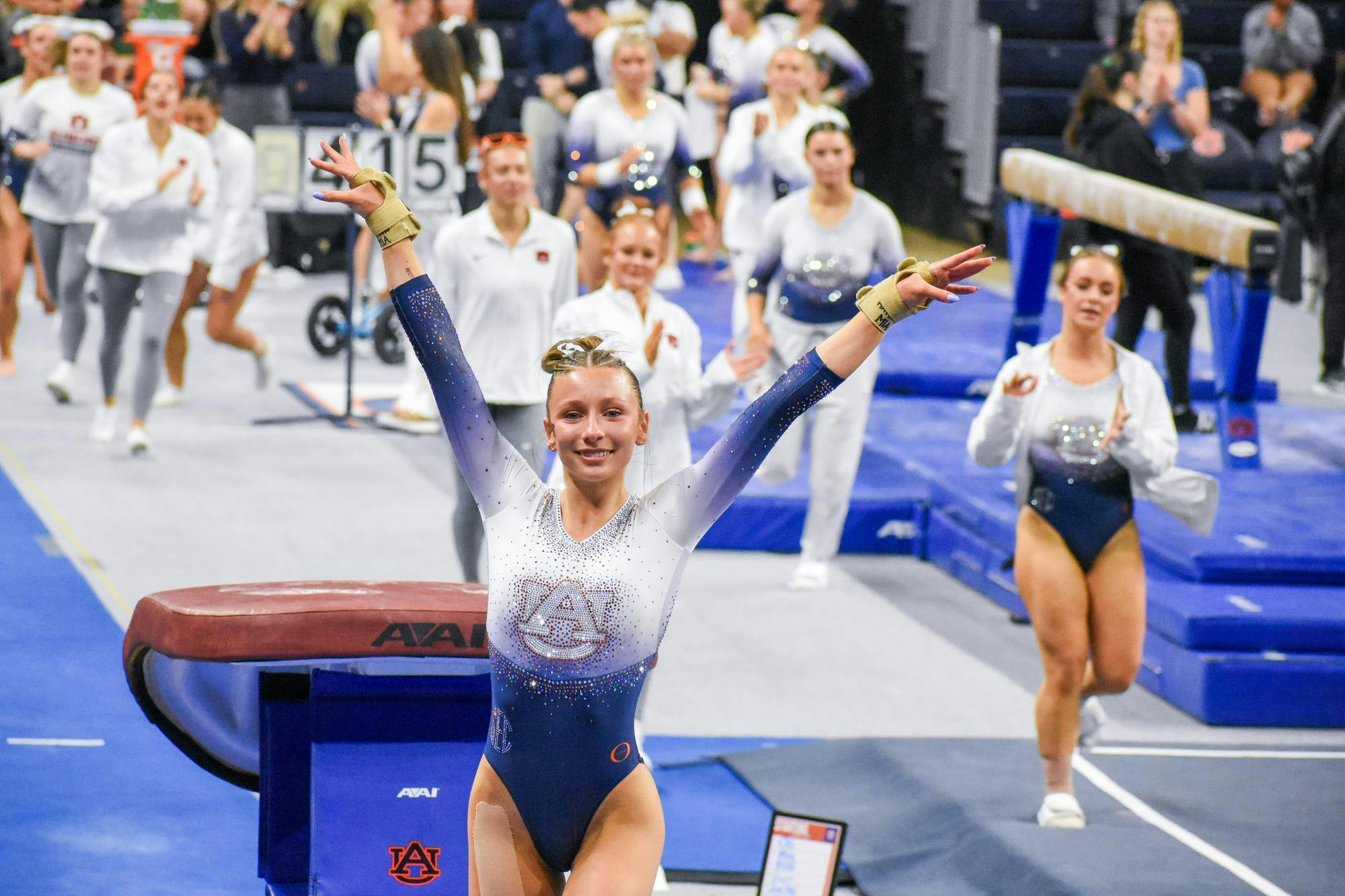 A gymnast in a sparkling leotard celebrates with her arms raised in front of a cheering crowd at a competition.