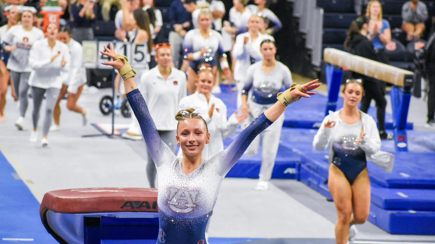 A gymnast in a sparkling leotard celebrates with her arms raised in front of a cheering crowd at a competition.