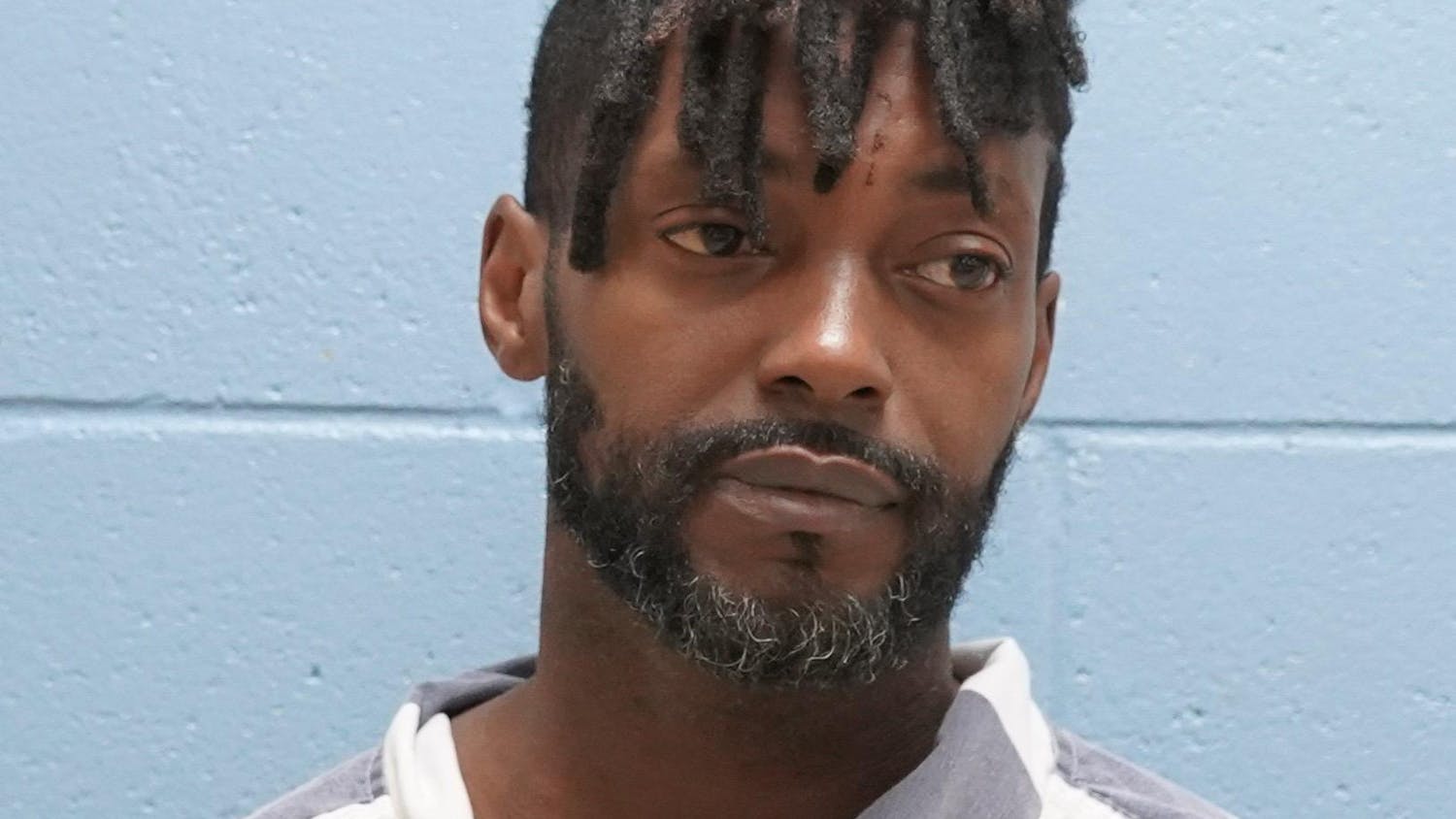 A man with a beard wears a striped prison uniform, standing against a light blue wall.