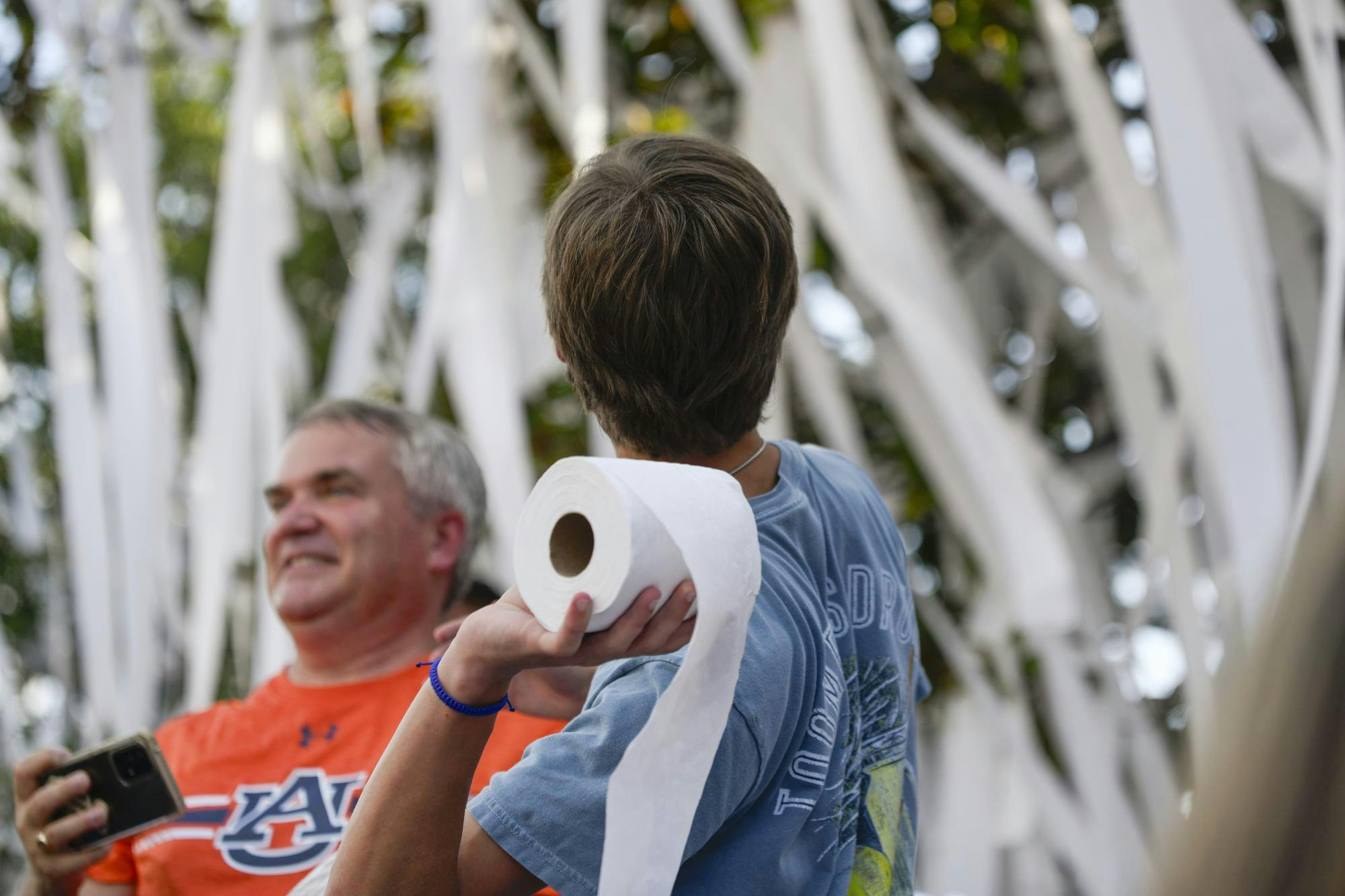 A young person holds a roll of toilet paper while a man smiles nearby, surrounded by strands of white material.