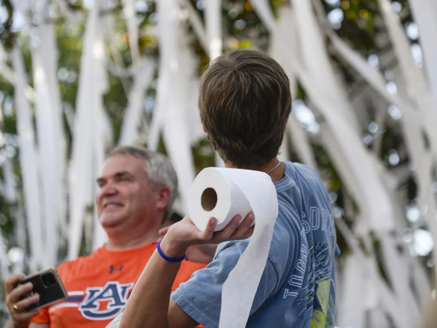 A young person holds a roll of toilet paper while a man smiles nearby, surrounded by strands of white material.