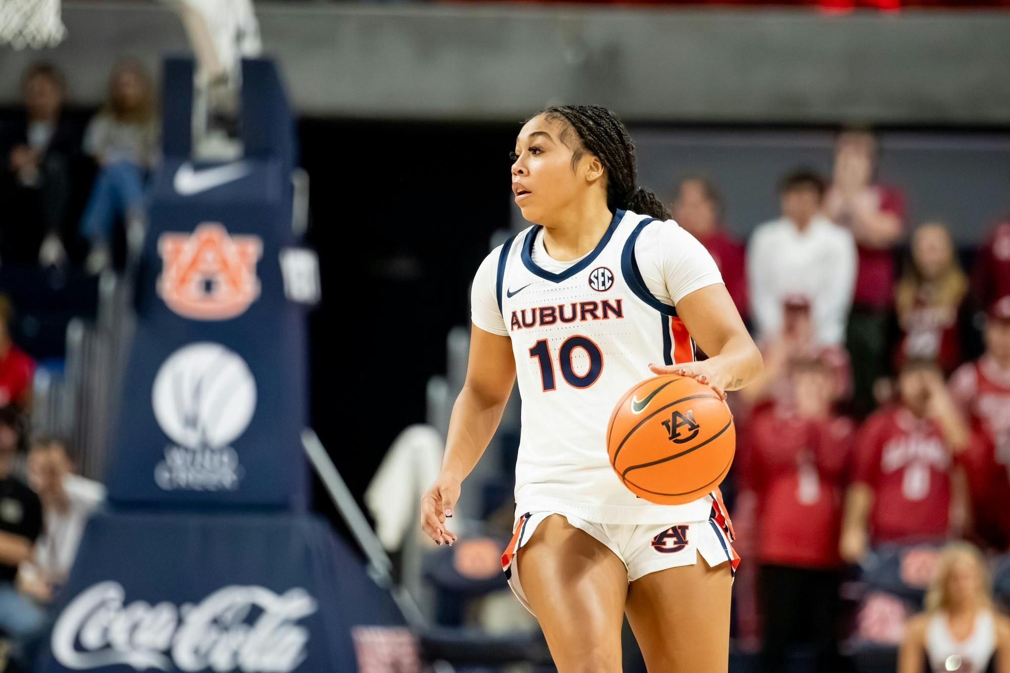 A female basketball player in a white and navy uniform dribbles a basketball while focused, with fans visible in the background.