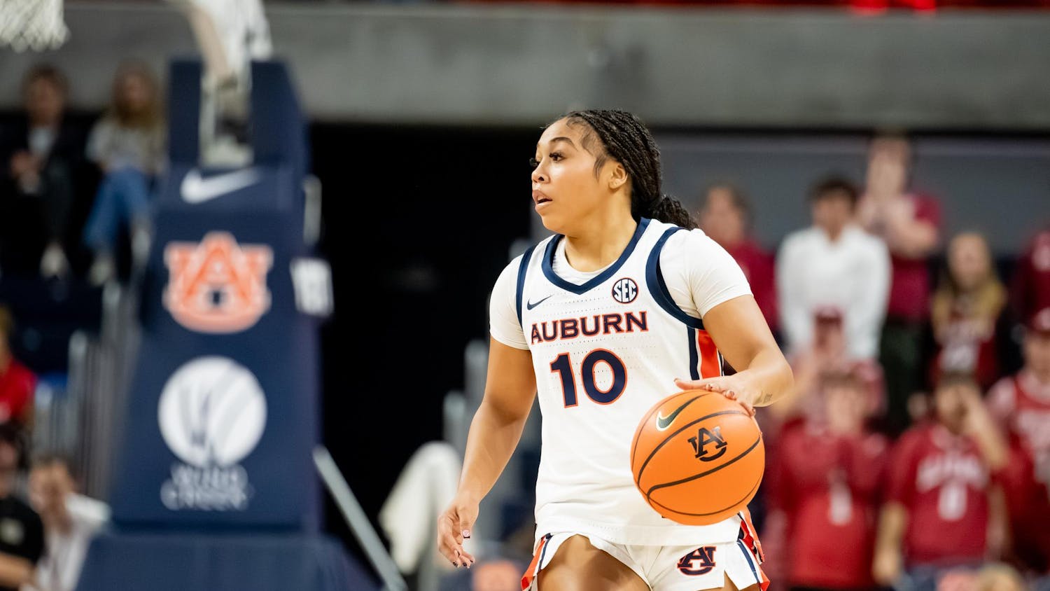 A female basketball player in a white and navy uniform dribbles a basketball while focused, with fans visible in the background.