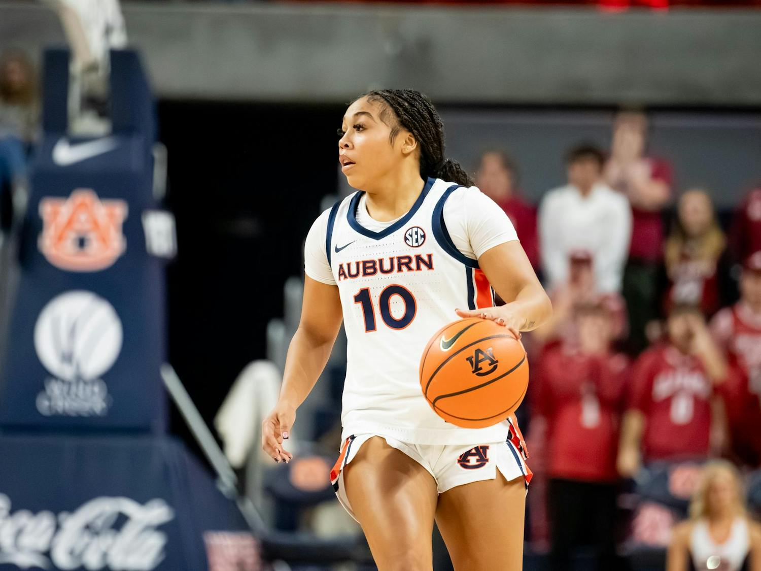 A female basketball player in a white and navy uniform dribbles a basketball while focused, with fans visible in the background.