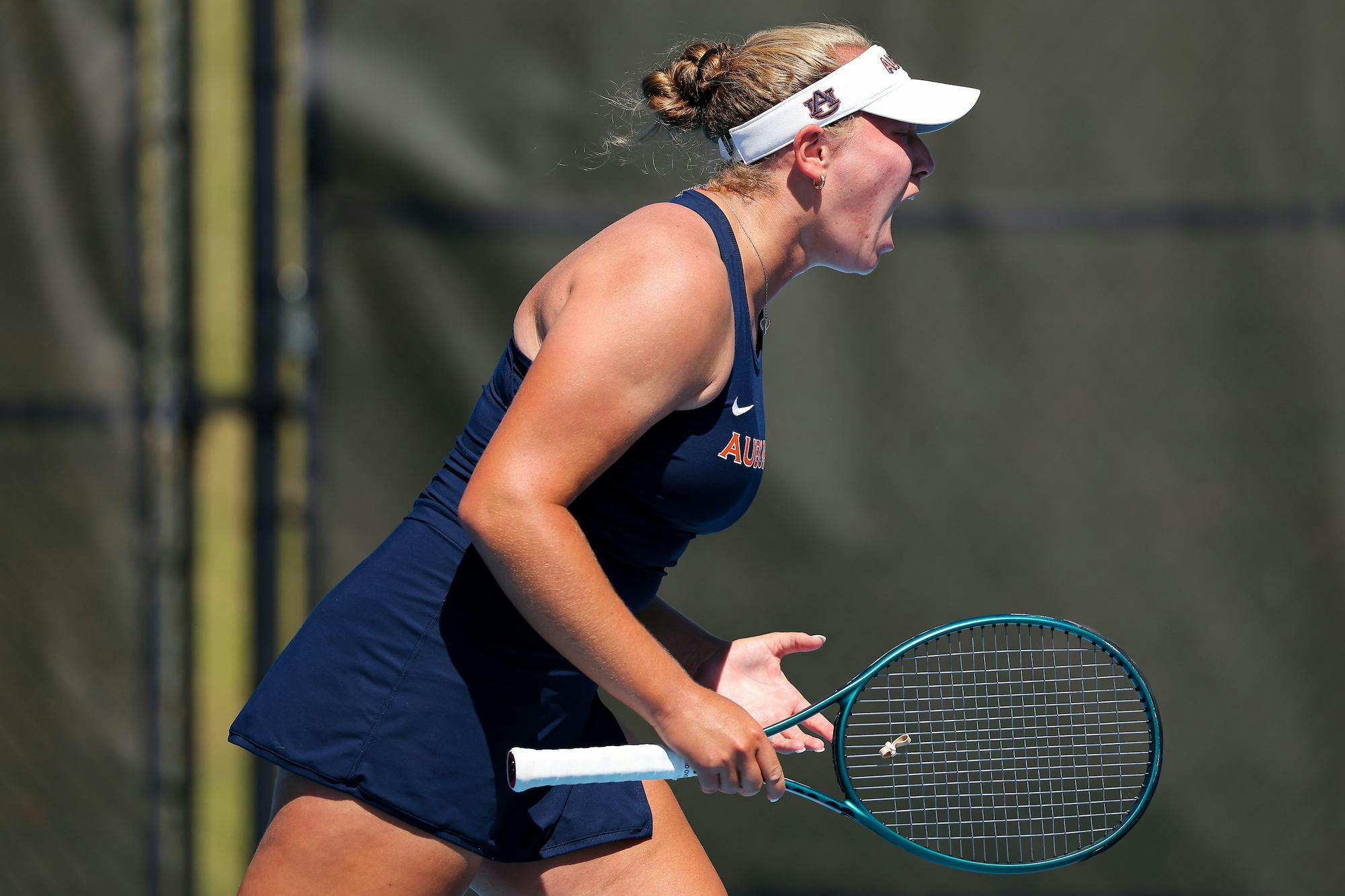 A young female tennis player, wearing a dark outfit and visor, celebrates passionately while holding a tennis racket.