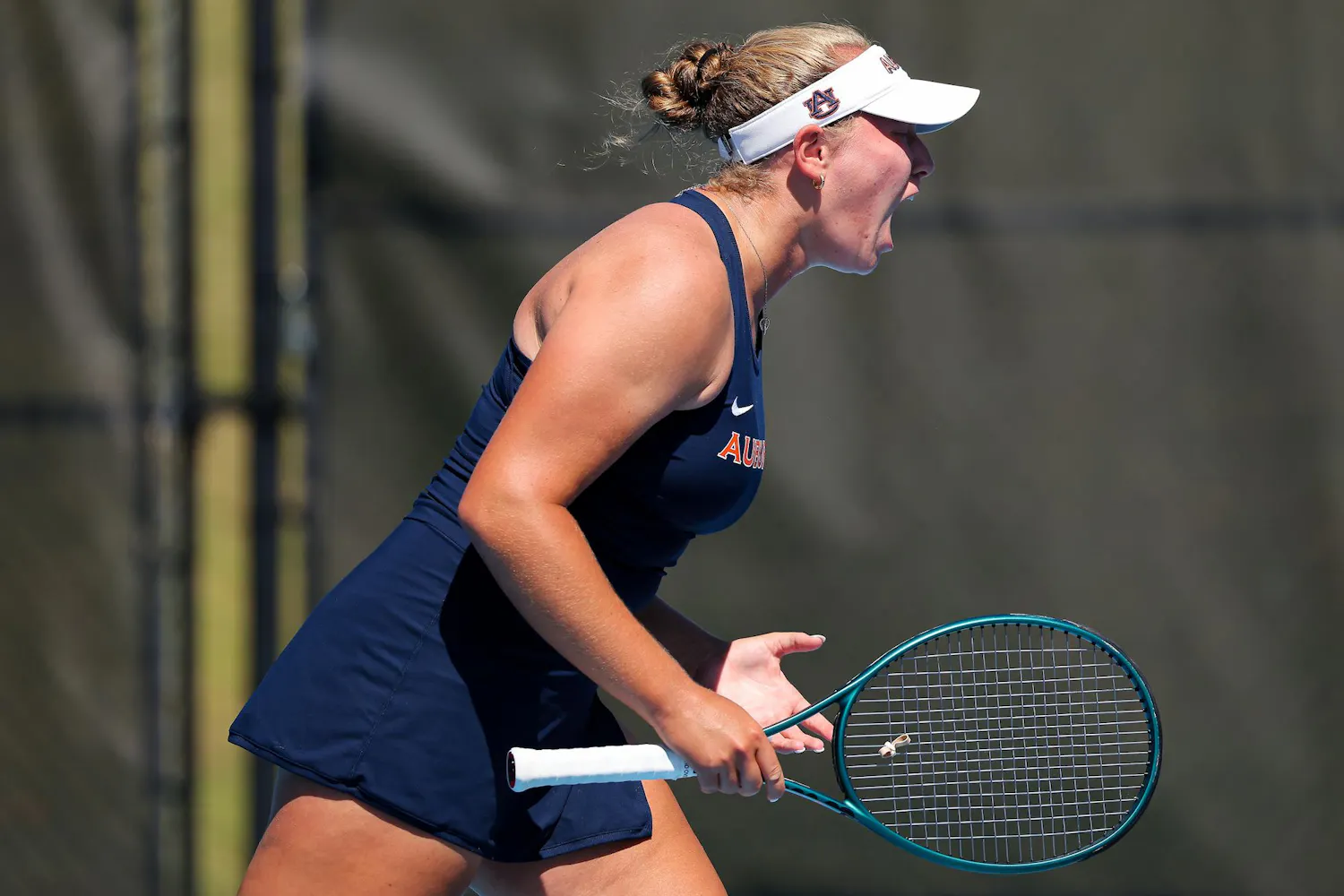 A young female tennis player, wearing a dark outfit and visor, celebrates passionately while holding a tennis racket.