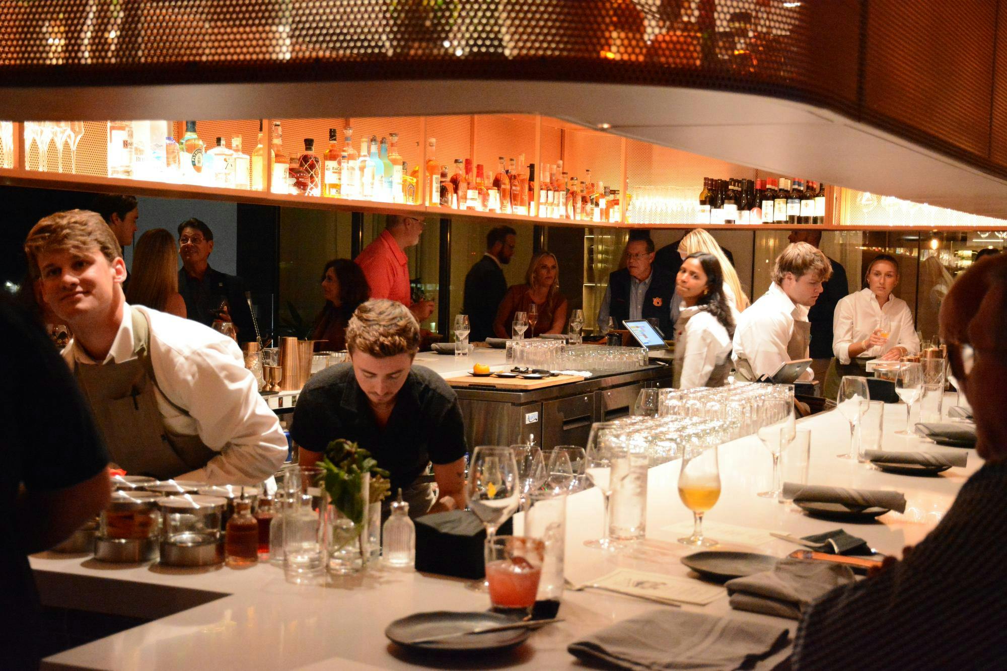 A bustling bar features staff in white shirts, guests mingling, and shelves lined with various bottles of liquor.