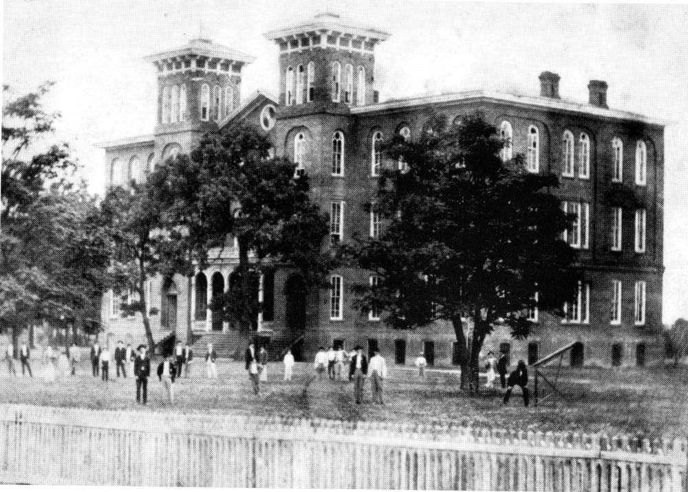 A large brick building with multiple towers stands behind a grassy area where several people in period clothing are gathered.