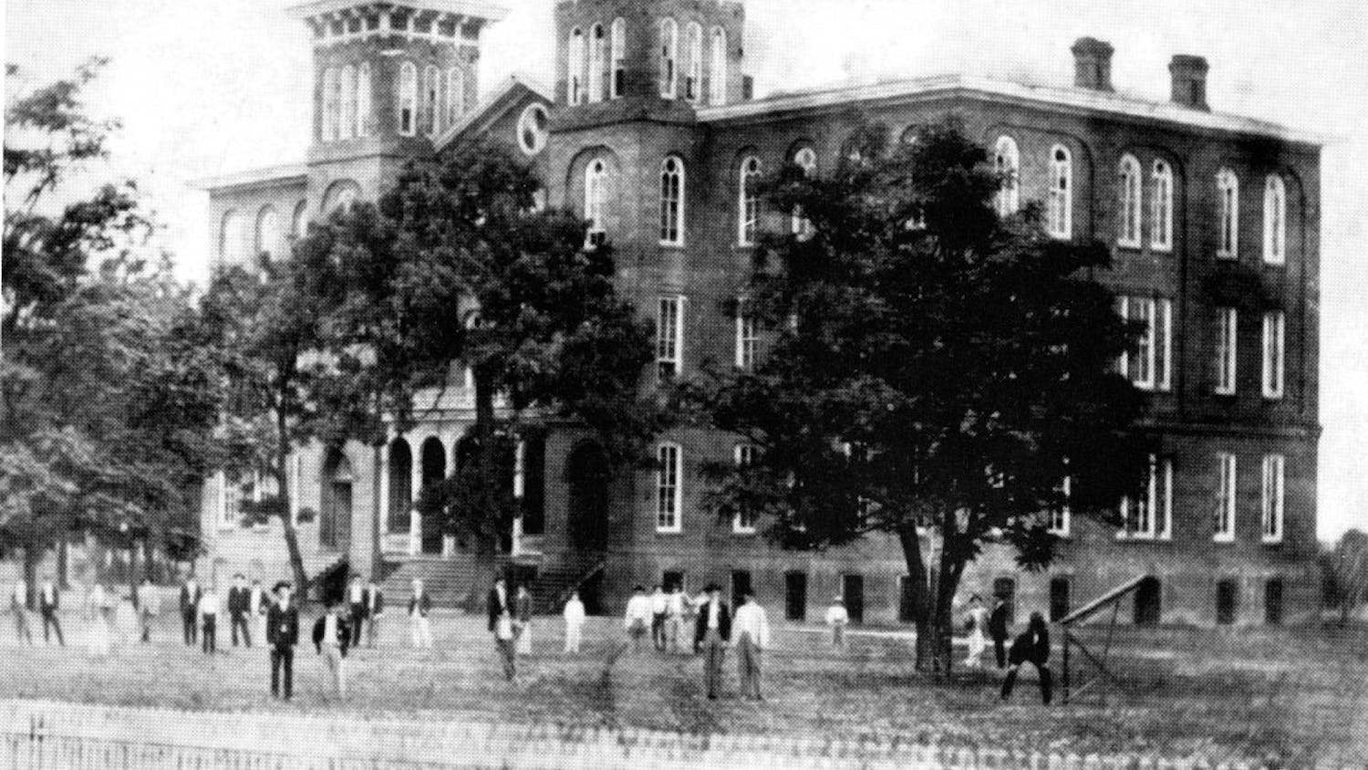 A large brick building with multiple towers stands behind a grassy area where several people in period clothing are gathered.