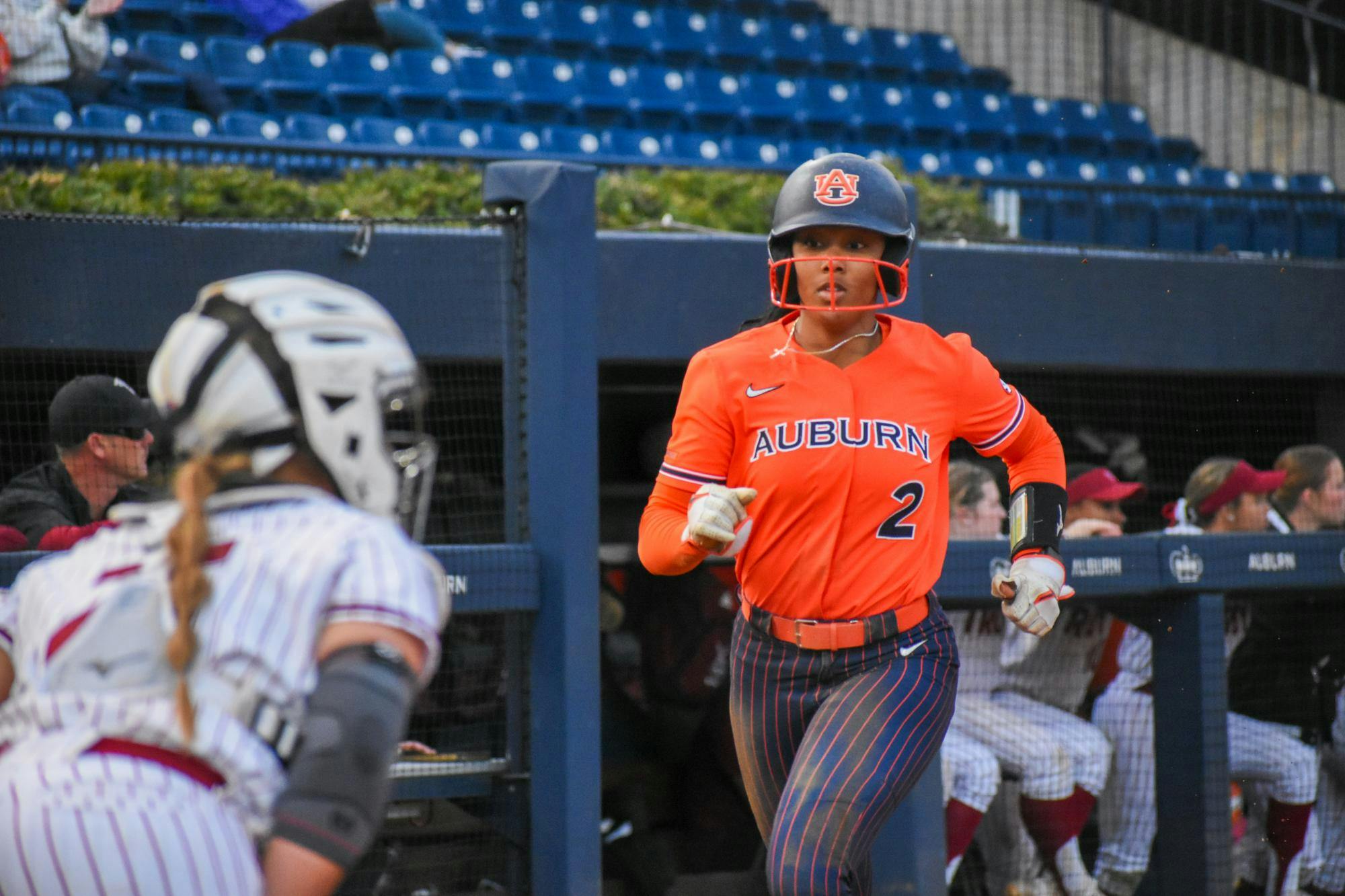 A softball player in an orange uniform with "AUBURN" written on it runs towards home plate, while a teammate in a striped outfit prepares to catch.