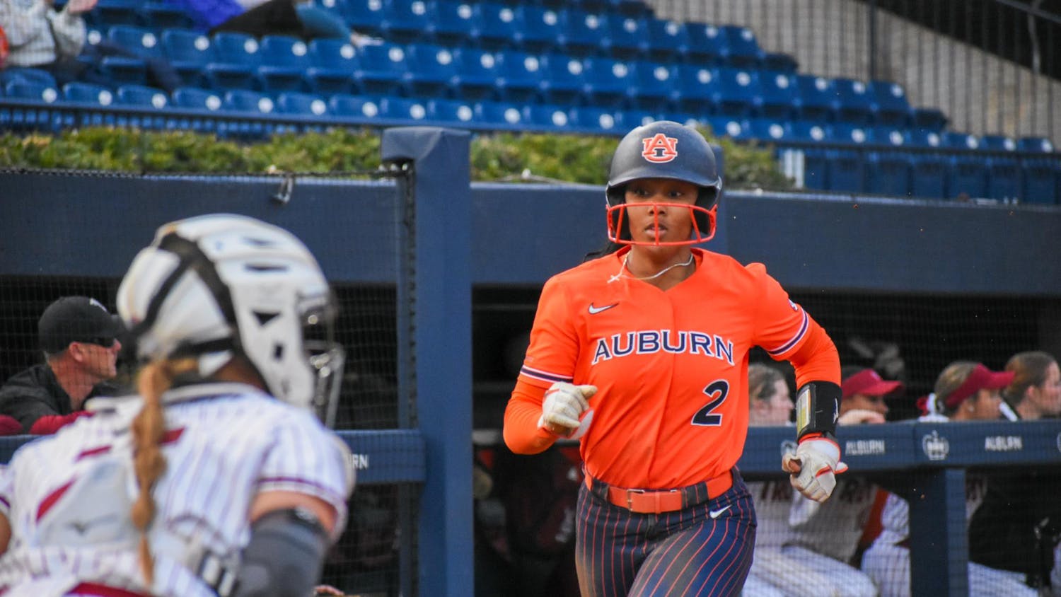 A softball player in an orange uniform with "AUBURN" written on it runs towards home plate, while a teammate in a striped outfit prepares to catch.