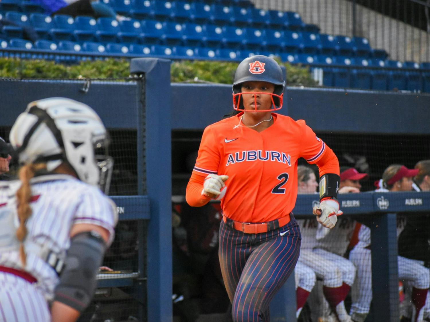 A softball player in an orange uniform with "AUBURN" written on it runs towards home plate, while a teammate in a striped outfit prepares to catch.
