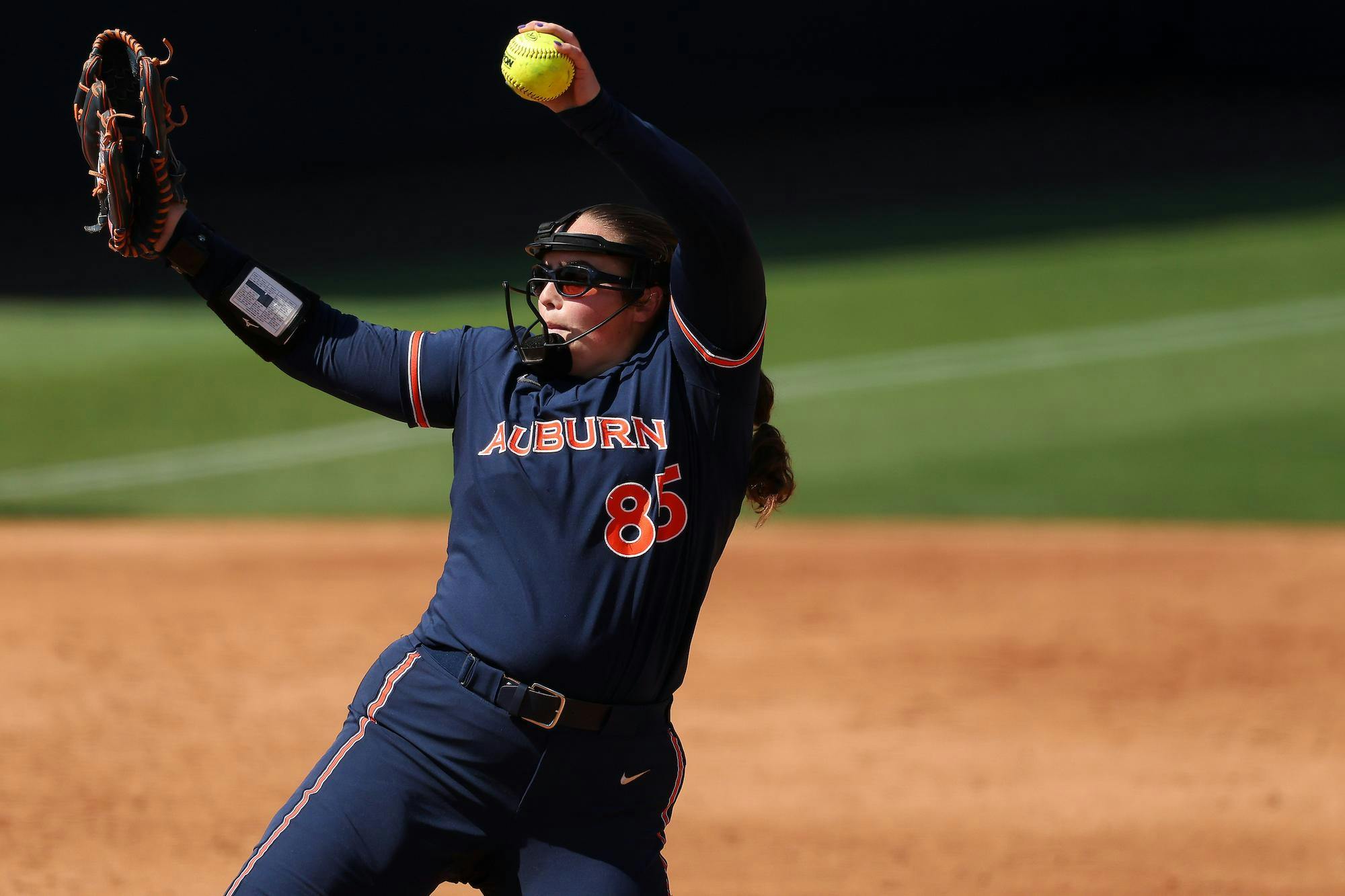 A young female softball player in a dark blue uniform winds up to pitch, holding a bright yellow softball in her right hand.
